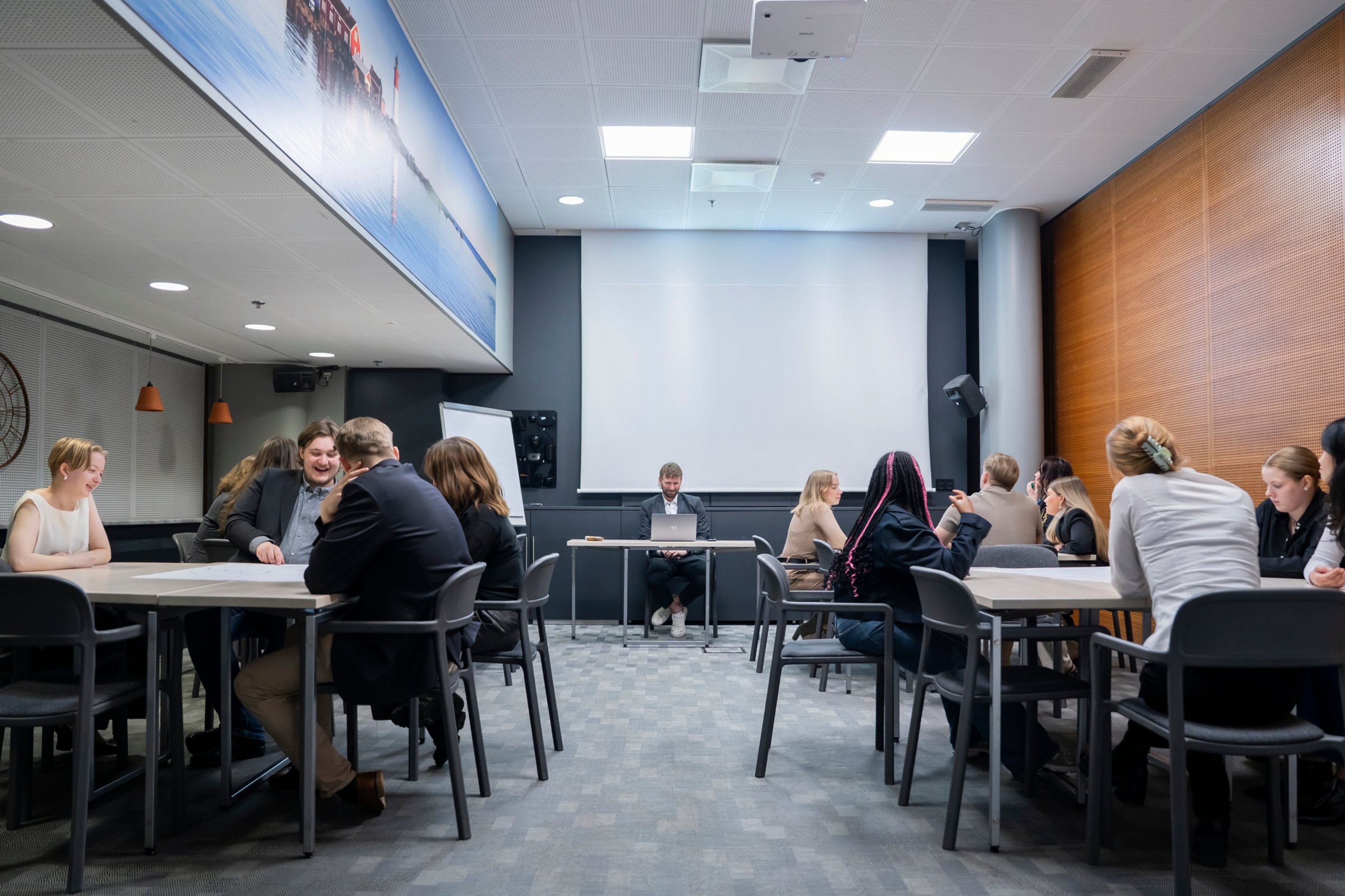 Moderni kokoushuone, jossa pöytiä ja tuoleja ryhmissä, valkokangas edessä ja puupaneelit sivuseinällä. Tasainen lattiapinta ja hyvä valaistus.