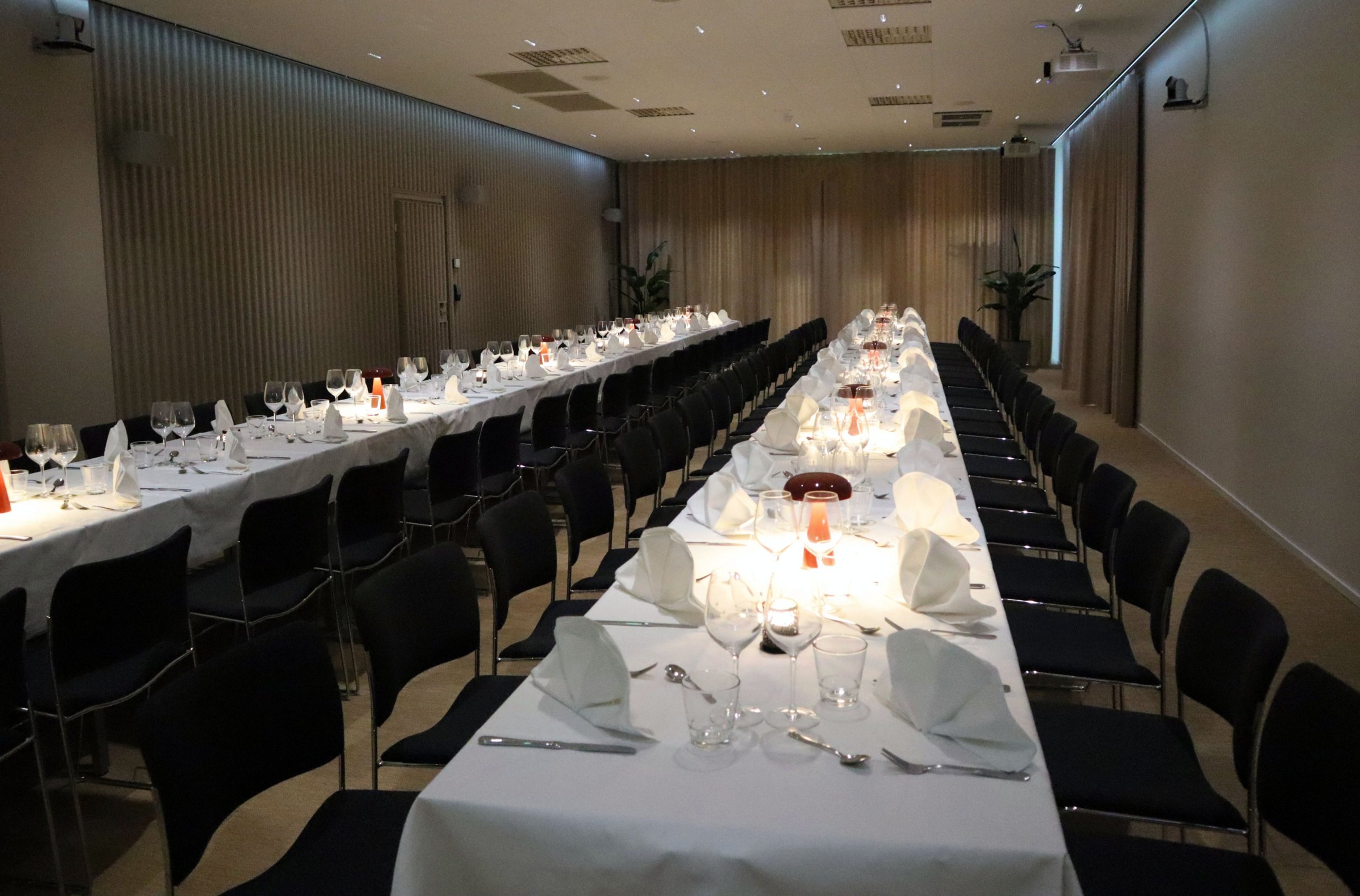 Kaksi pitkää juhlapöytää mustilla tuoleilla varustettuna, valkoliinaisina ja kattauksella. Tila on avara, tasalattiainen ja hyvin valaistu.