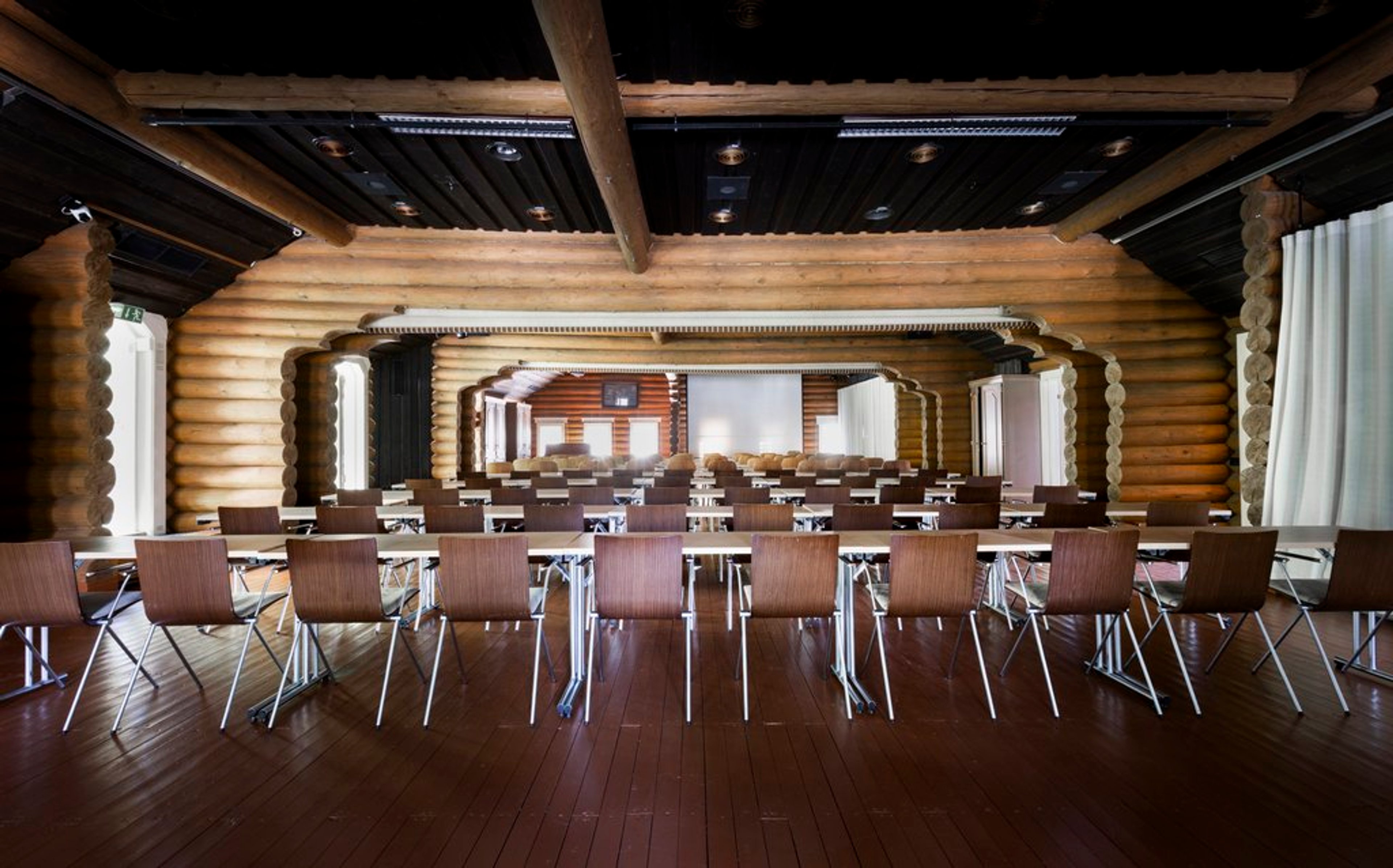 Conference venue with log timber walls, glossy wooden floor and rows of brown modern chairs. White projection screen at the back.