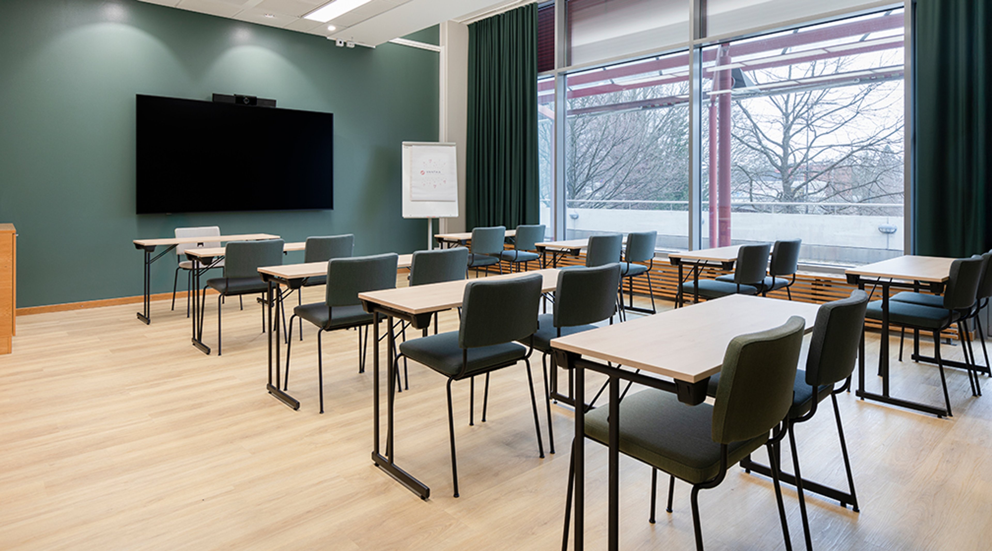 Moderni kokoushuone vihreällä seinällä, vaalealla puulattialla ja pöytäryhmillä. Suurista ikkunoista näkyy puita, tilassa on näyttö seinällä.