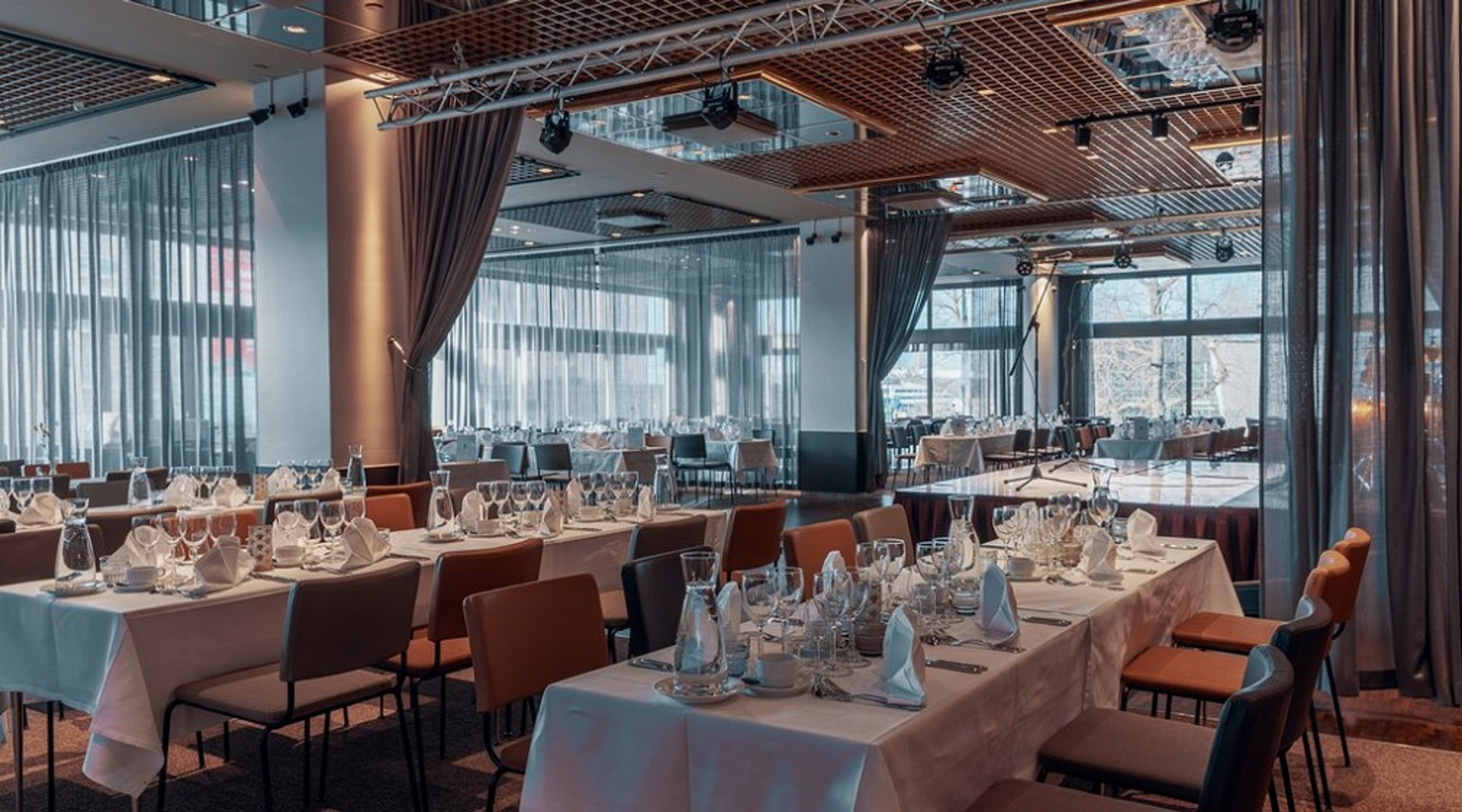 Moderni ravintolatila valkoliinaisilla pöydillä ja oransseilla tuoleilla. Sisustuksessa lasiseinät, harmaat verhot ja puiset kattoelementit.