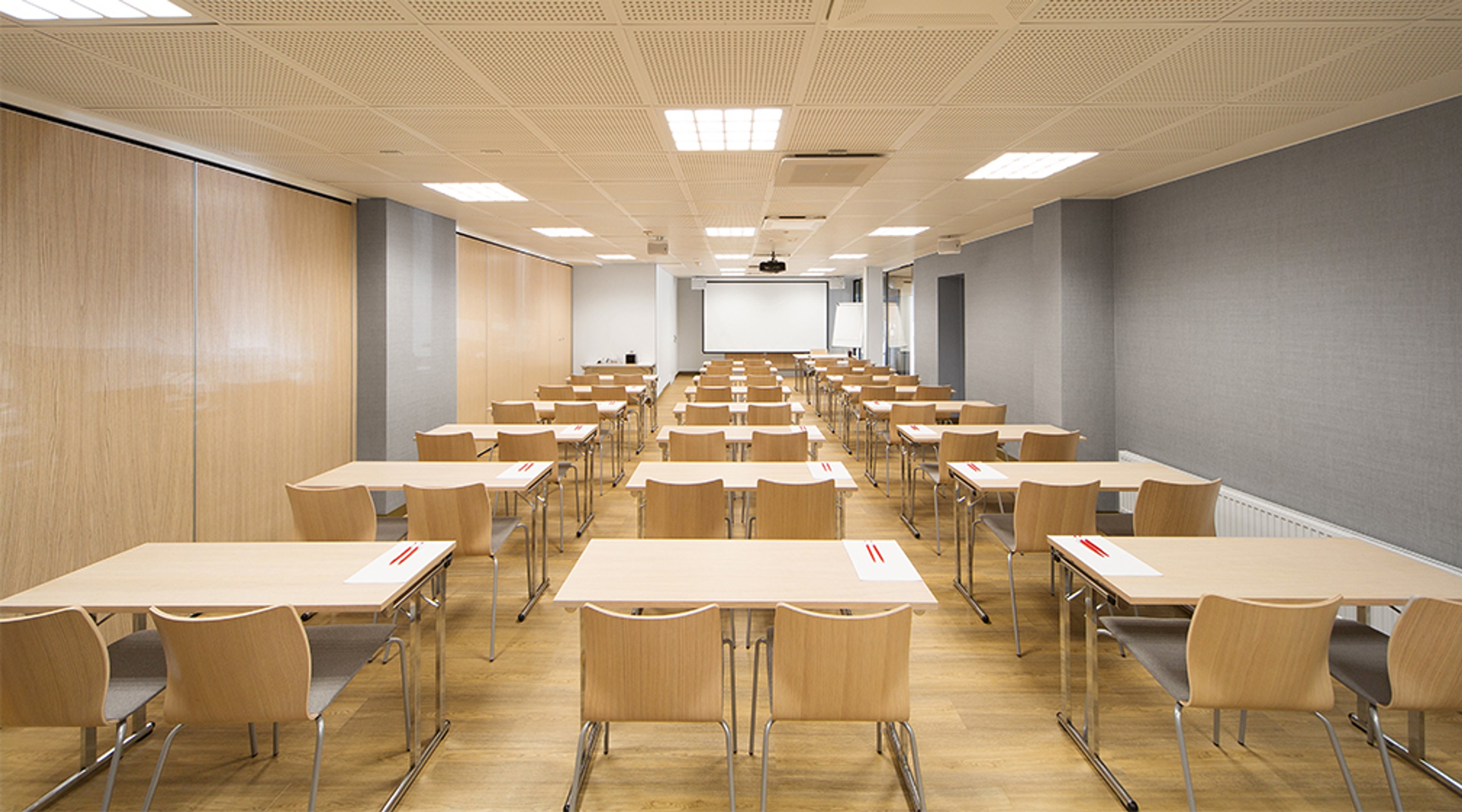 Moderni kokoushuone puulattialla, jossa useita pöytiä ja tuoleja riveittäin. Valkea projektiokangas edessä ja tasainen valaistus.