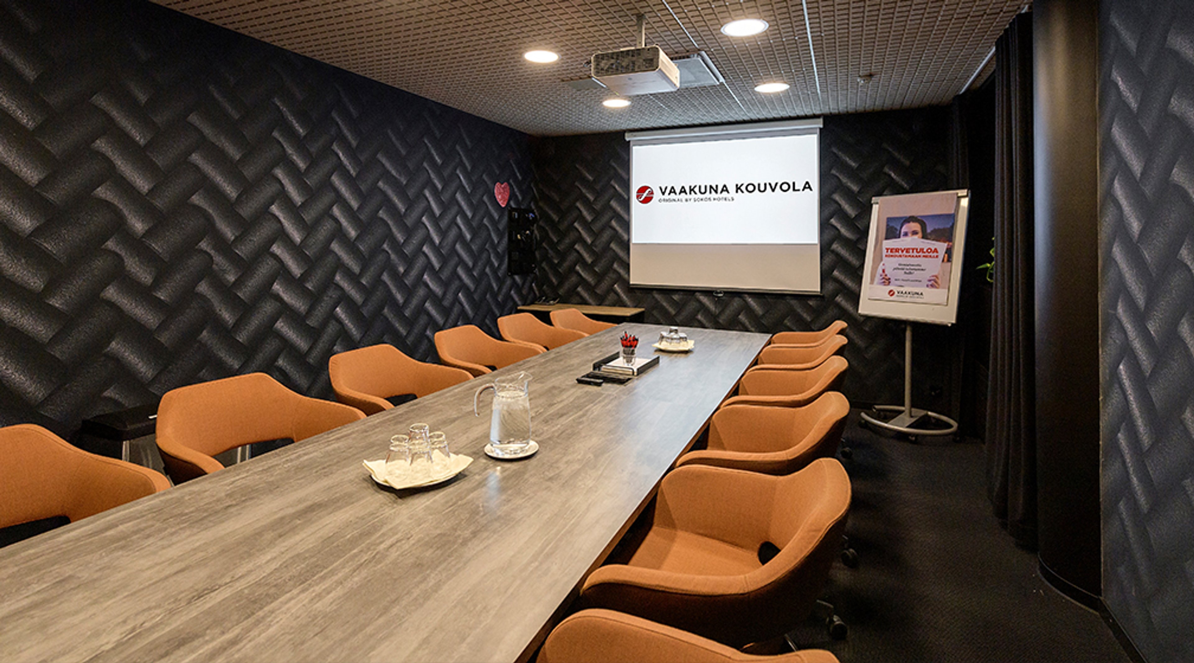 Moderni neuvotteluhuone teksturoiduilla mustilla seinillä, pitkä puinen pöytä ja oranssit tuolit. Valkokangas edessä, tilassa hyvä valaistus.