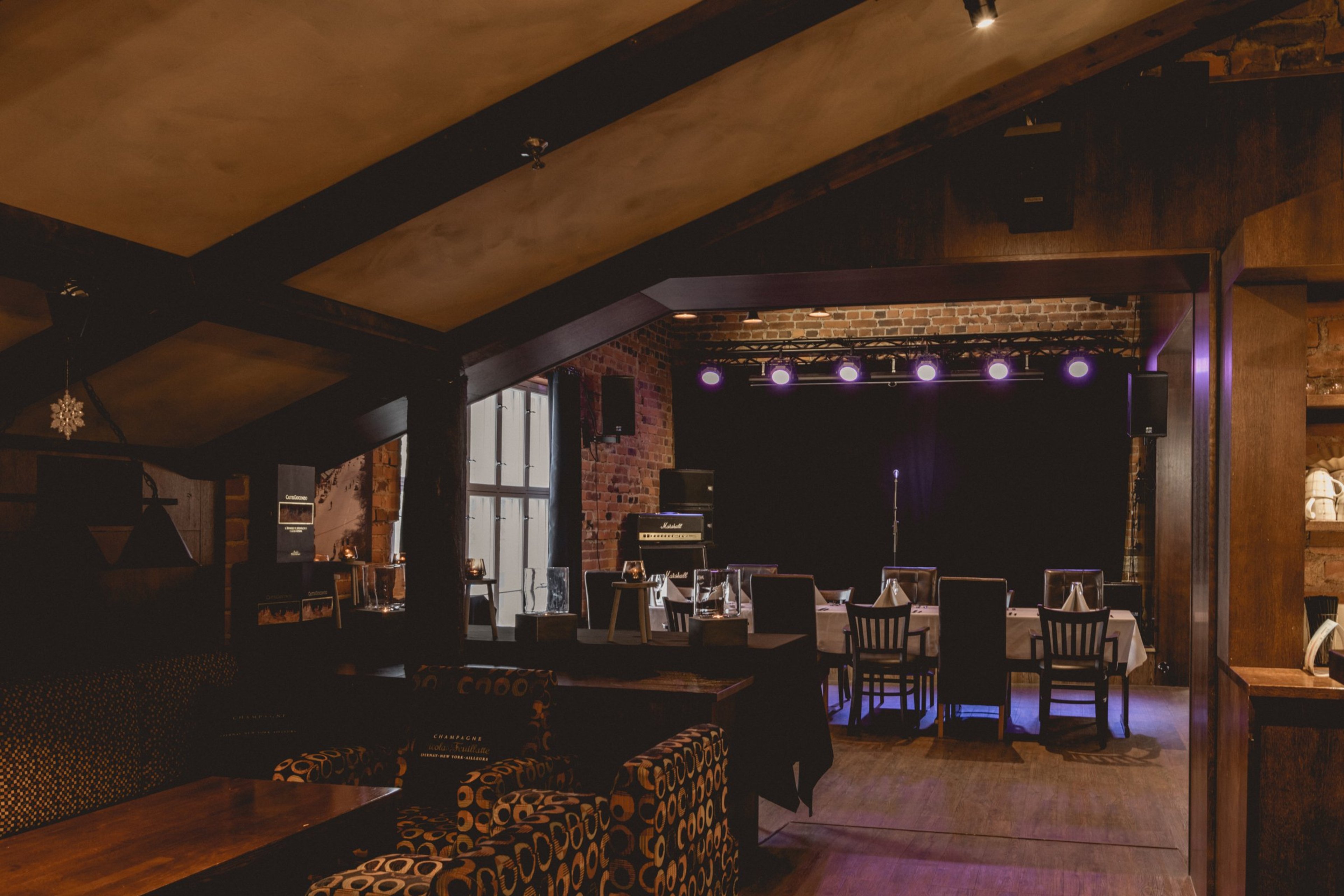 Ravintolasali, jossa tiiliseinäinen esiintymislava purppuroilla valoilla ja ruokapöytiä. Puulattia, kattohirret ja lounge-istuintila etualalla.