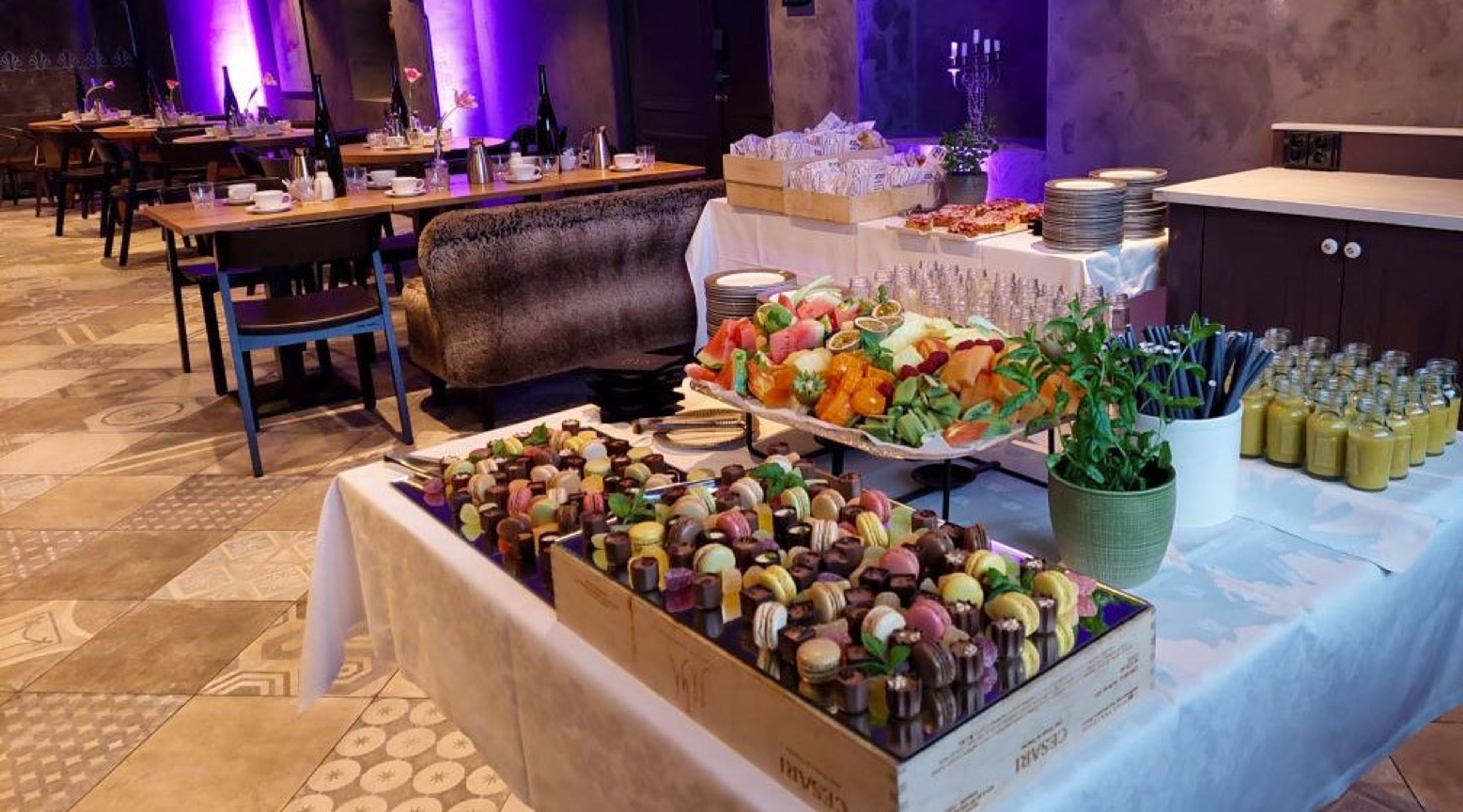 Tarjoilupöytä täynnä macaroneja, voileipälautasia ja smoothie-pulloja juhlatilassa, jossa purppura valaistus ja ravintolapöytiä taustalla.