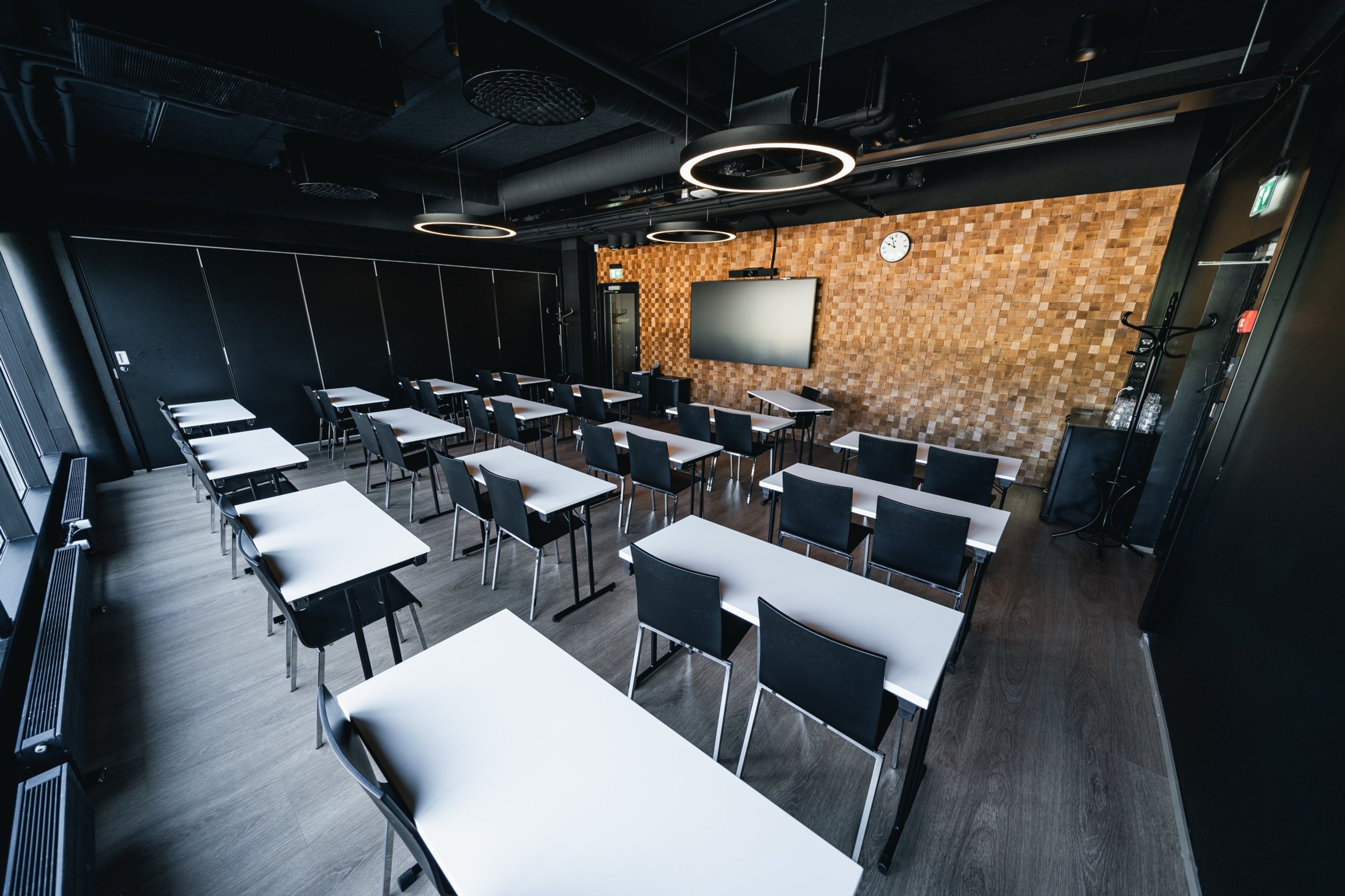 Moderni luokkahuone valkoisilla pöydillä ja mustilla tuoleilla. Tilassa on puinen seinä ja näyttö. Lattia on kovaa puuta ja valaistus on rengasmaisilla kattovalaisimilla.