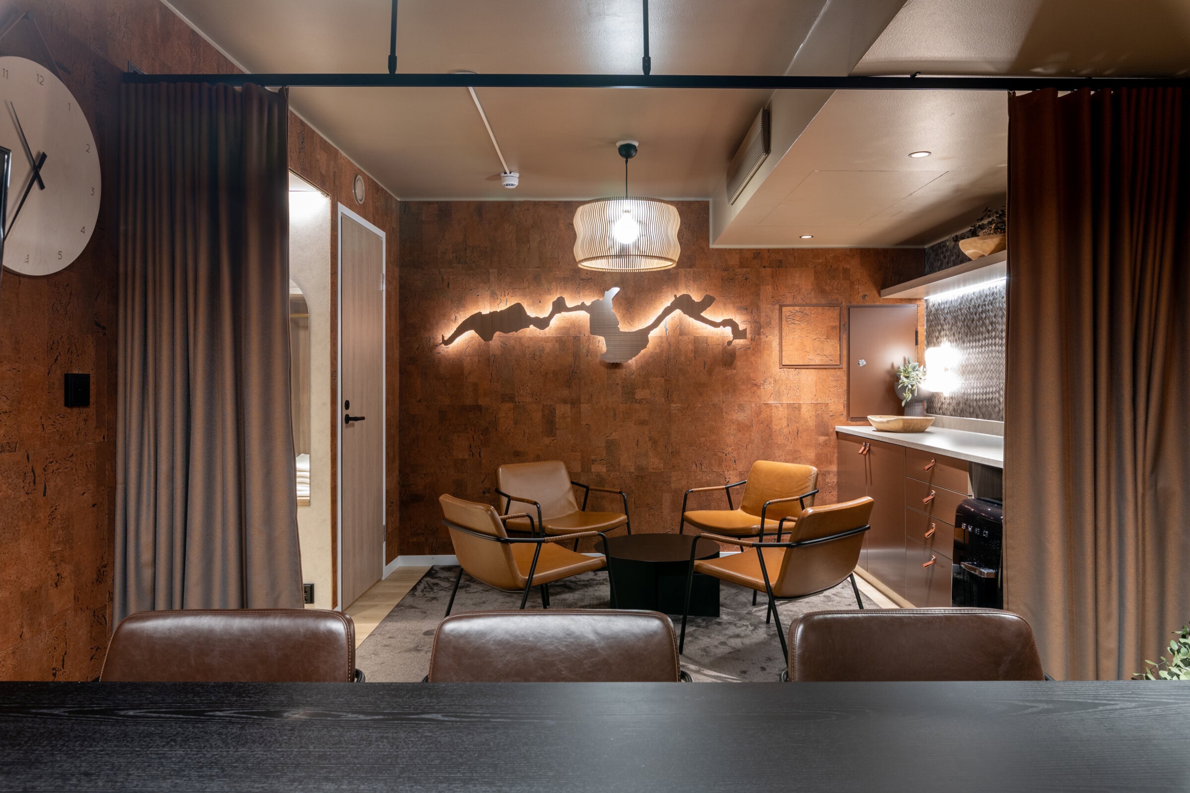 Living room with cork walls, four-seat seating area behind green curtain dividers and kitchenette. Concrete-coloured floor.