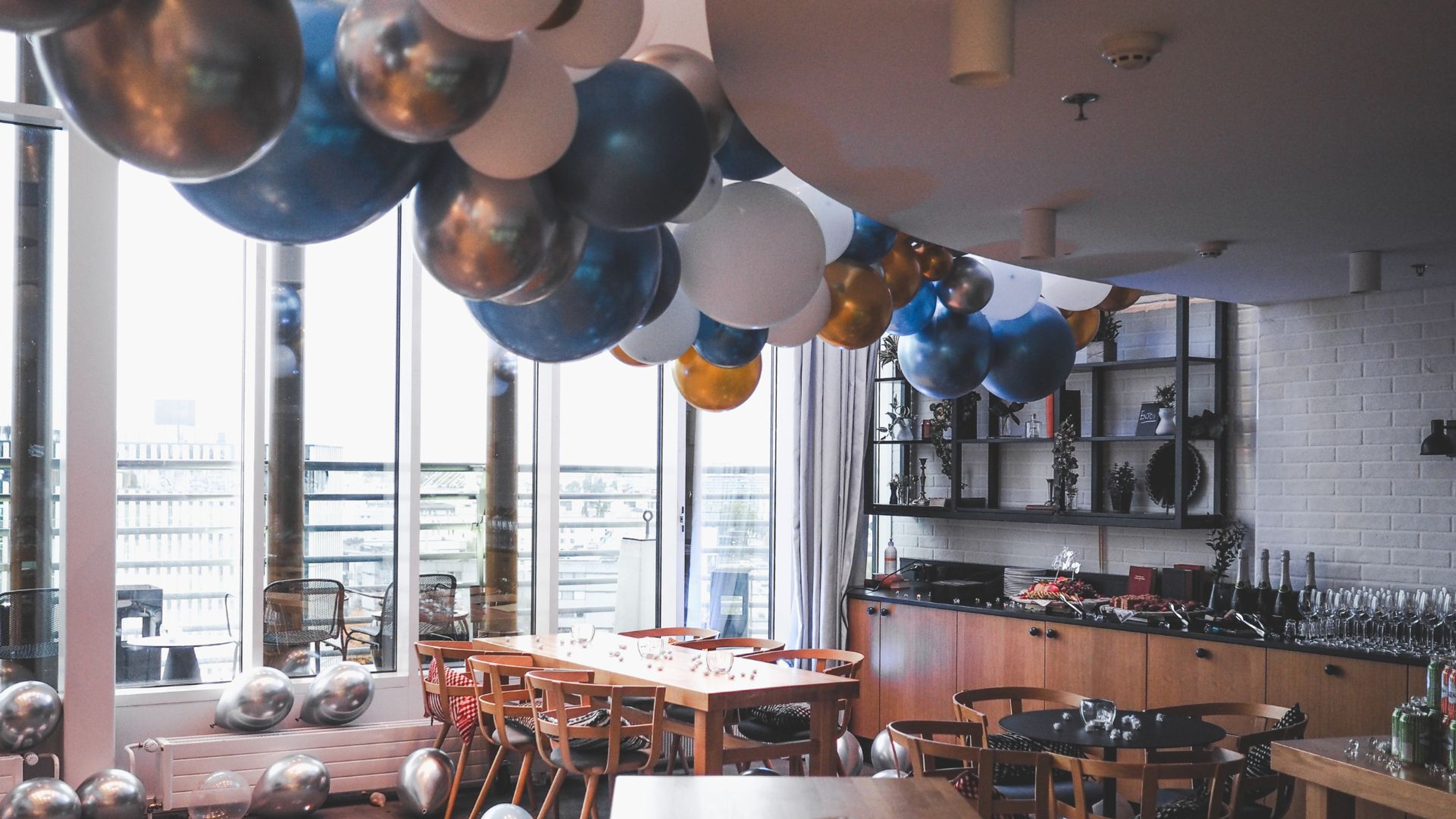 Juhlava tila koristeltu sinisillä, valkoisilla ja kullanvärisillä ilmapalloilla. Tilassa puiset pöydät, suuri ikkuna ja tarjoilupiste.