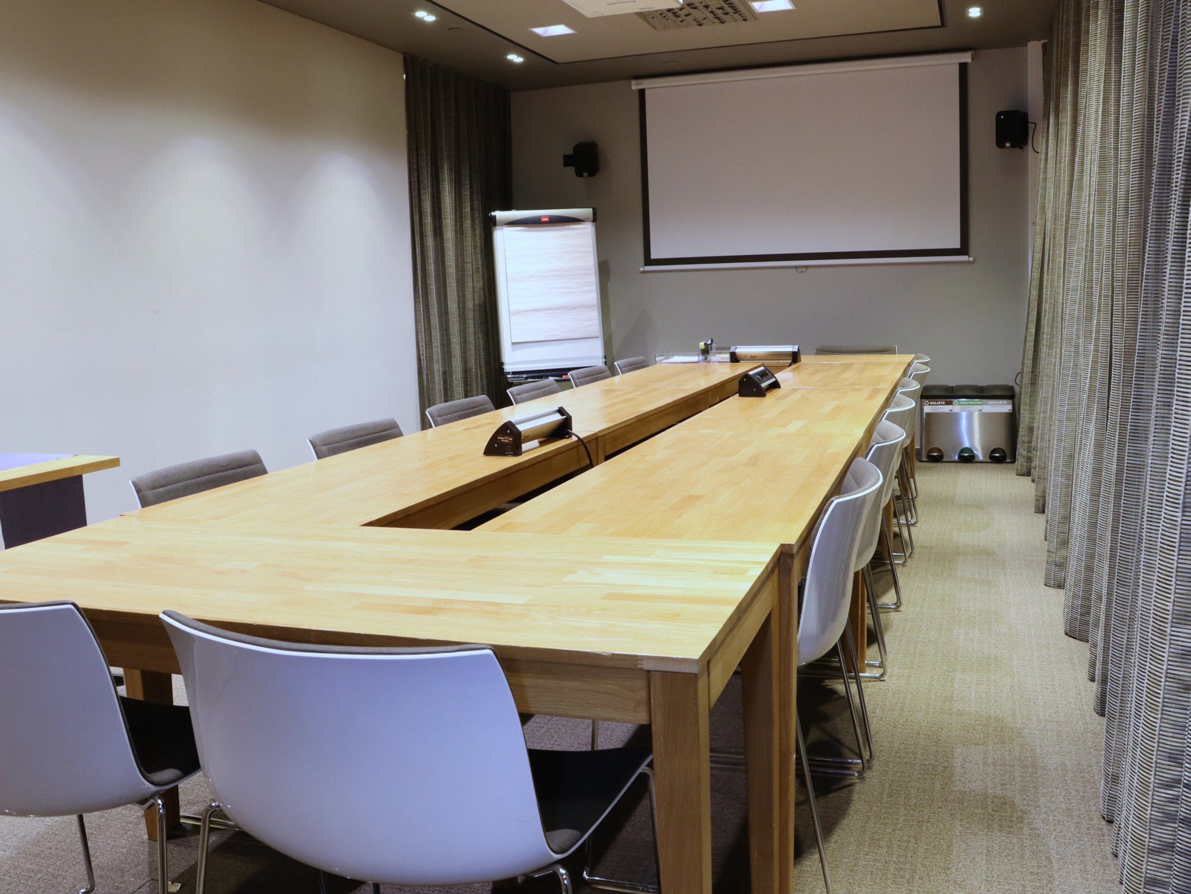 Moderni kokoushuone U-muotoisella puupöydällä, valkoisilla tuoleilla, esityskankaalla ja fläppitaululla. Tasainen lattia ja hyvä valaistus.