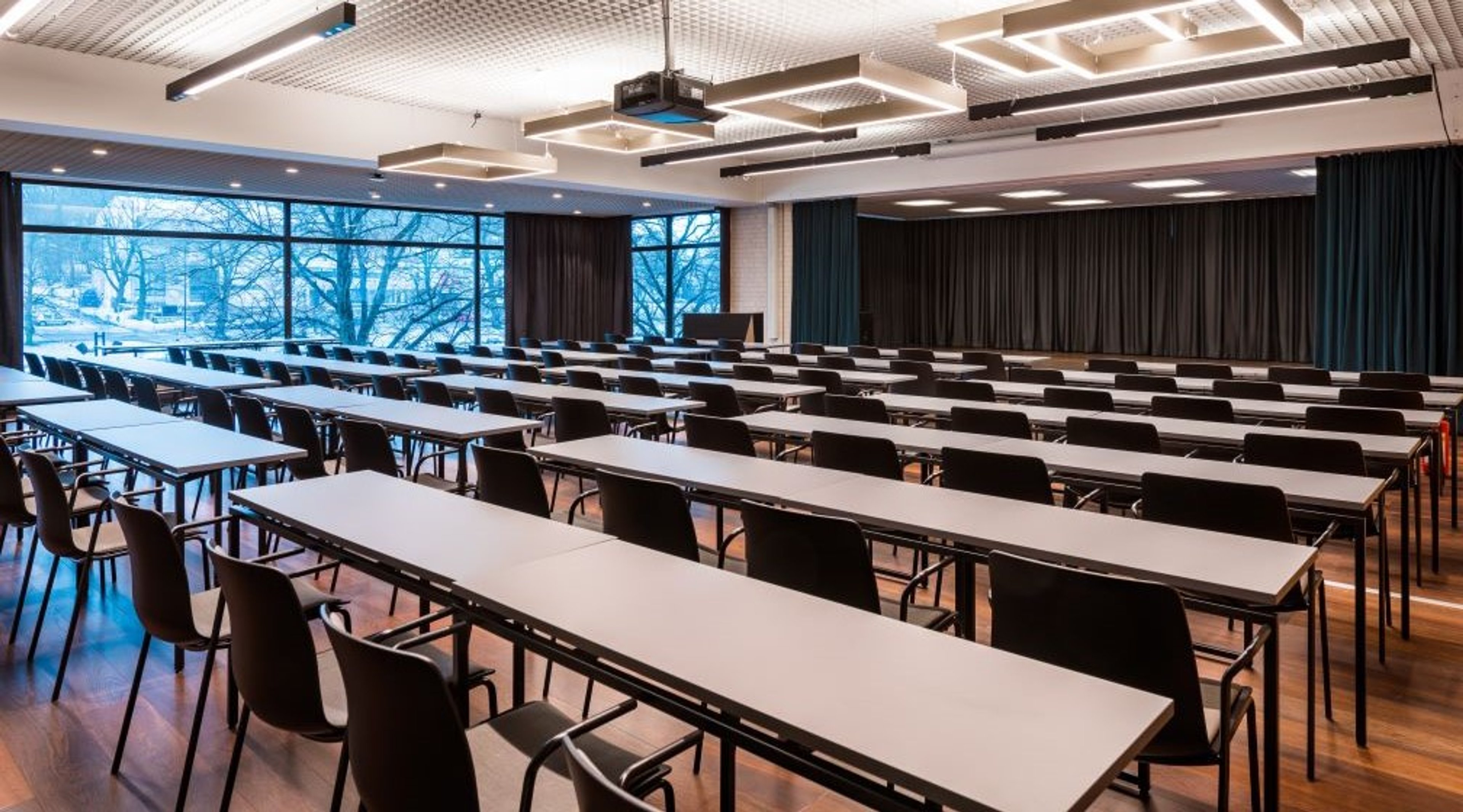 Moderni auditorio pitkillä pöydillä ja tuoleilla, isot ikkunat näyttävät talvimaisemaan, puulattia ja tummat verhot.