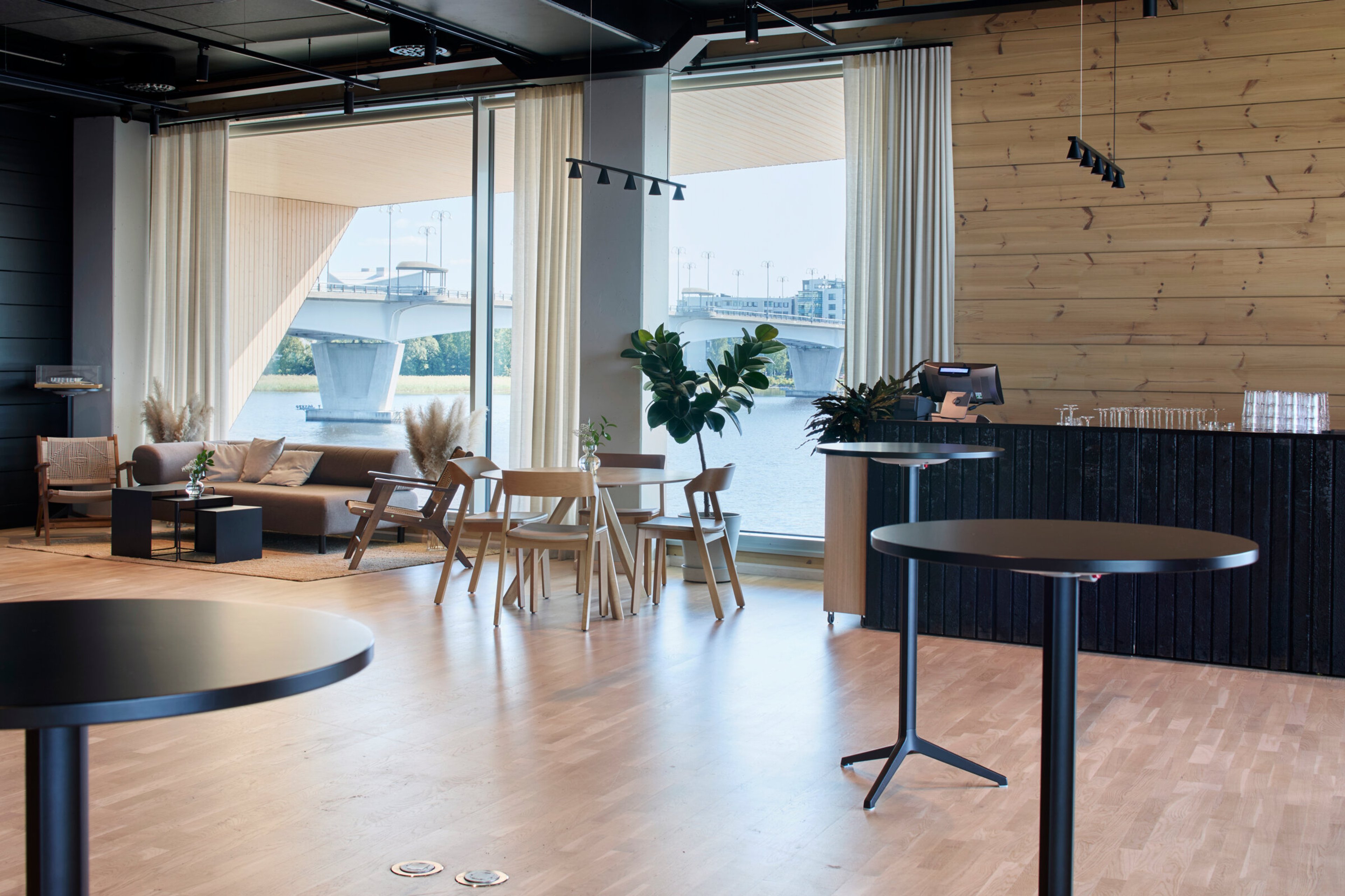 Moderni kahvilatila puulattialla. Suurista ikkunoista näkyy silta ja vettä. Lounge-alue sohvalla, pöytiä, tuoleja ja baaritiski. Esteetön tasainen lattia.