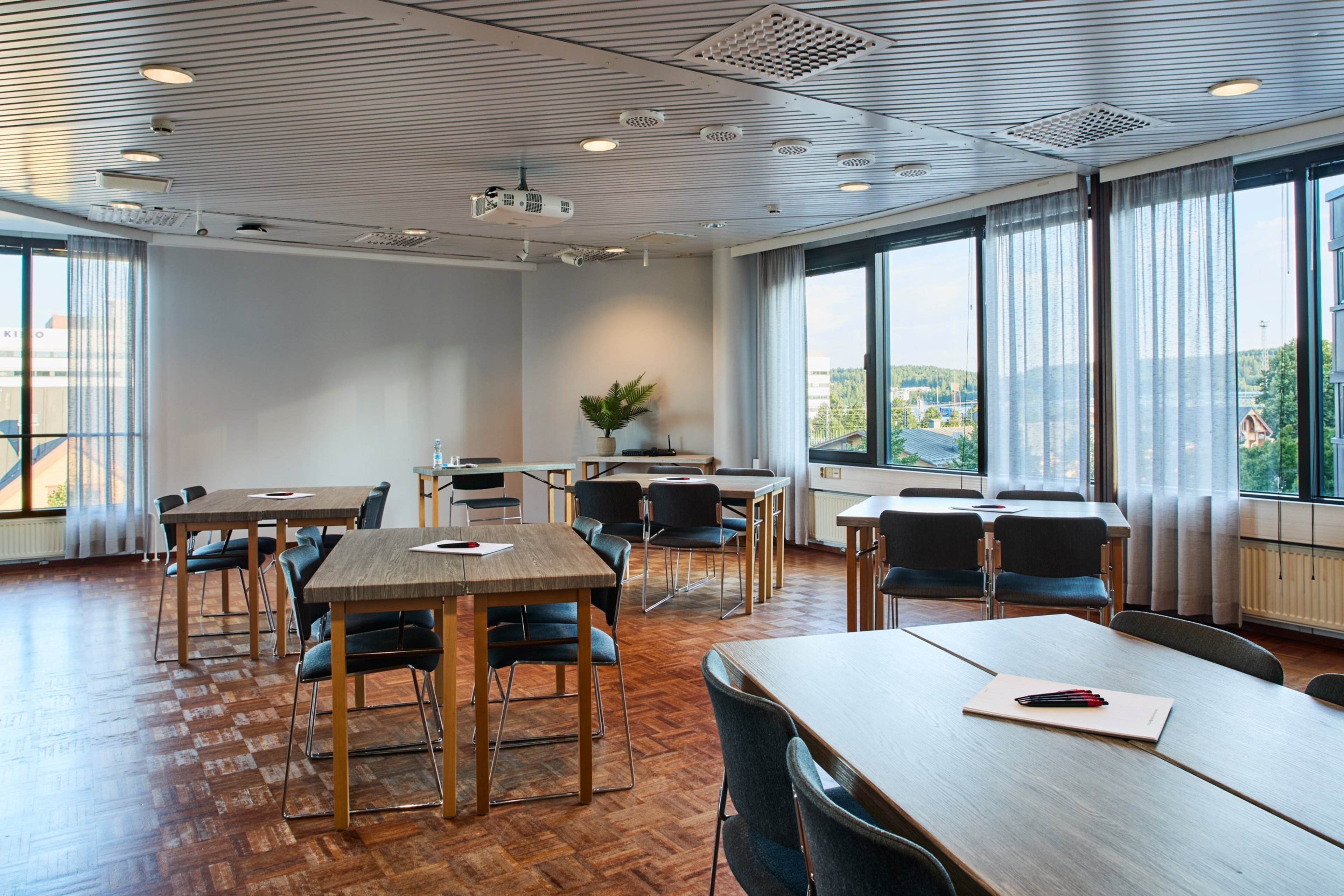 Valoisa kokoustila puulattialla, jossa on useita pöytiä ja tuoleja. Suurista ikkunoista avautuu näkymä ulos. Katossa on videotykki.