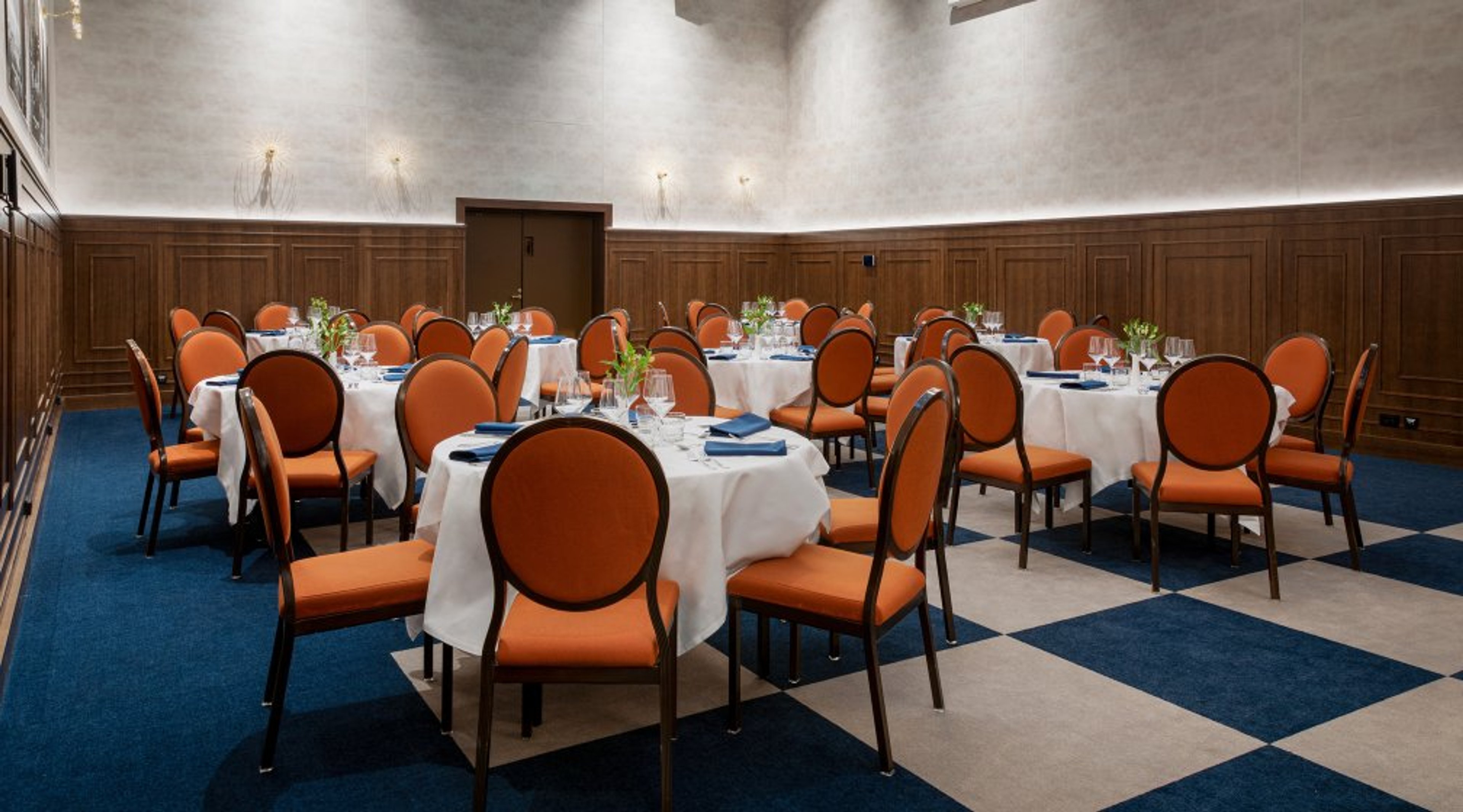 Ravintolasali, jossa on pyöreitä pöytiä valkoisilla liinoilla ja oransseilla tuoleilla. Lattialla sinivalkoinen ruutukuvio.