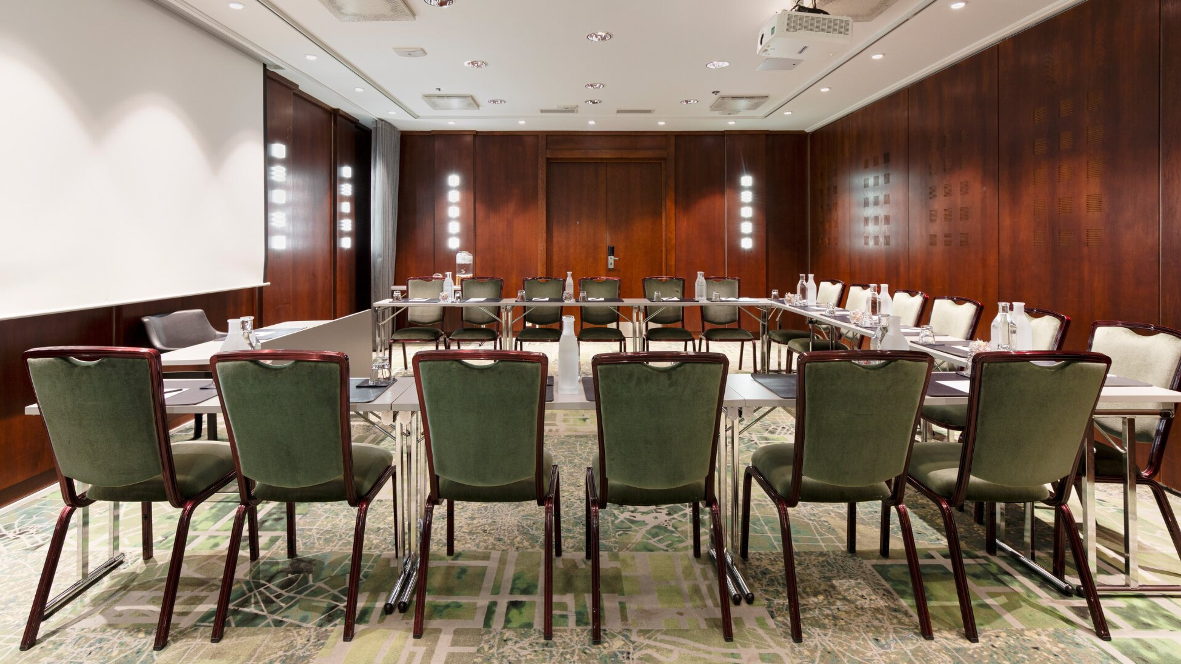 Kokoushuone puupaneeliseinillä ja vihreillä tuoleilla. Pöydät U-muodossa, valkokangas edessä. Tasainen lattiapinta, hyvä valaistus.