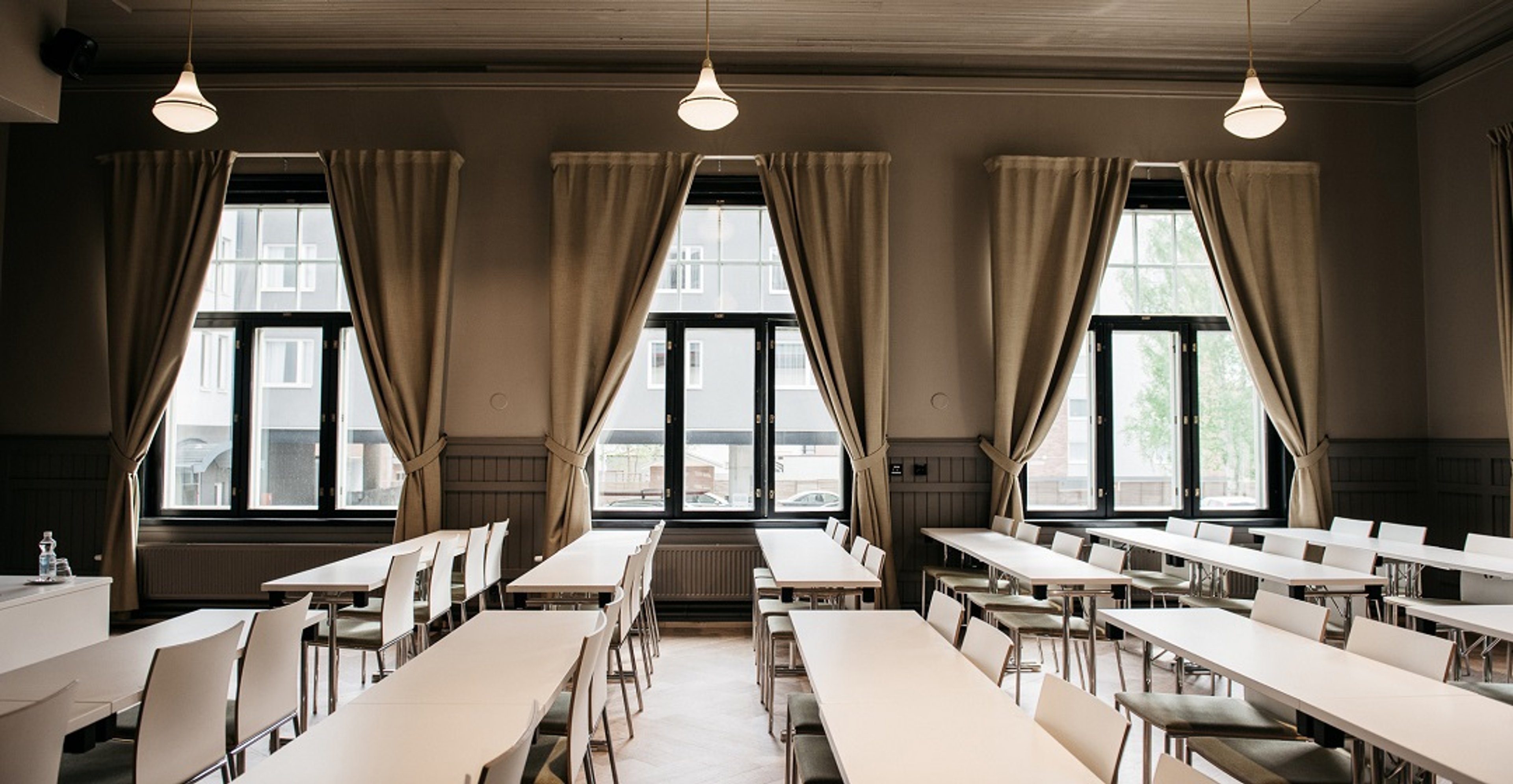 Kokoushuone, jossa pitkiä pöytiä ja valkoisia tuoleja. Suuret ikkunat beigein verhoin, kattovalaisimet luovat rauhallisen tunnelman.