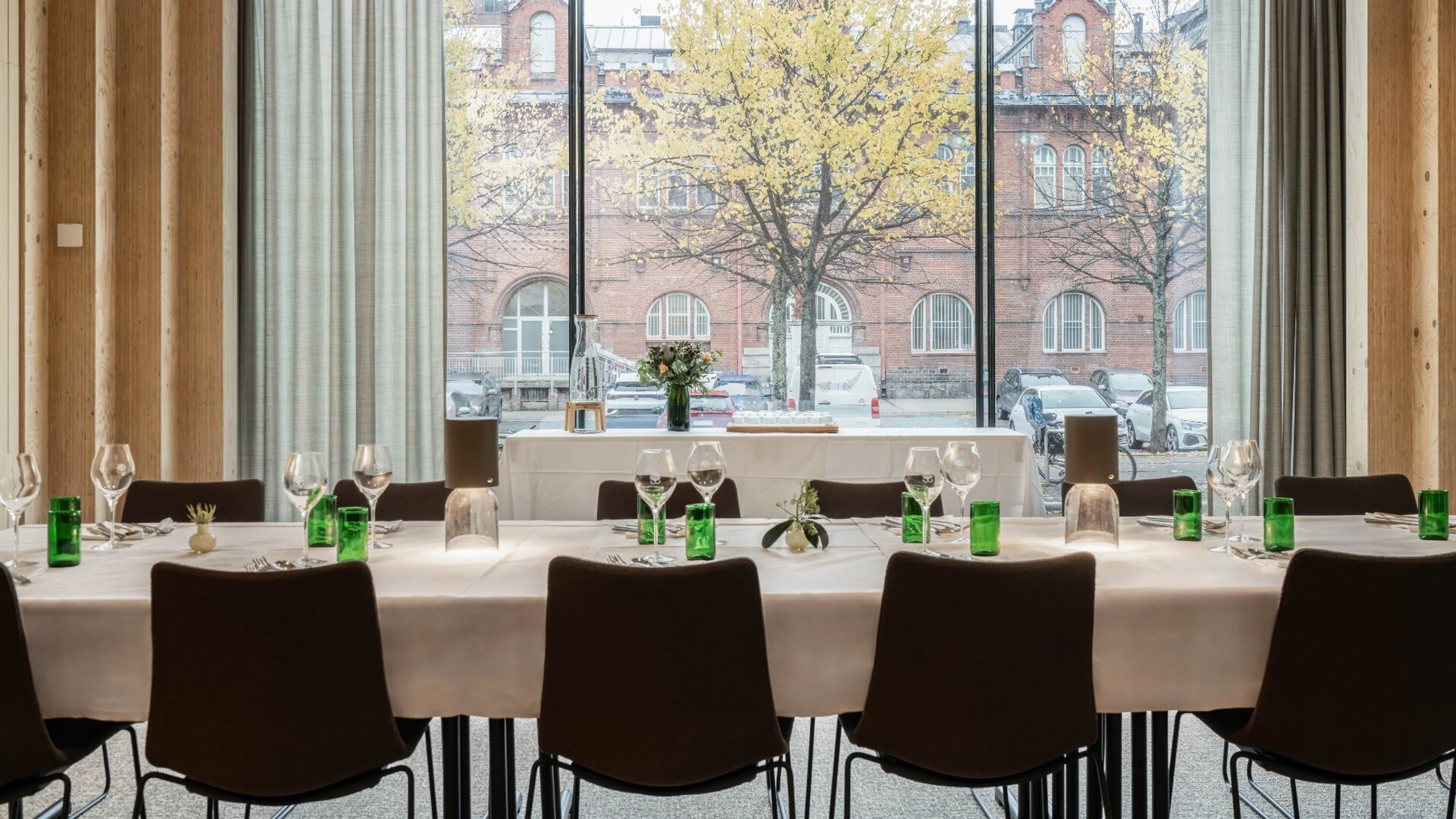 Restaurangbord med glas och stolar, genom stora fönster syns en tegelbyggnad och ett träd med gula höstlöv.