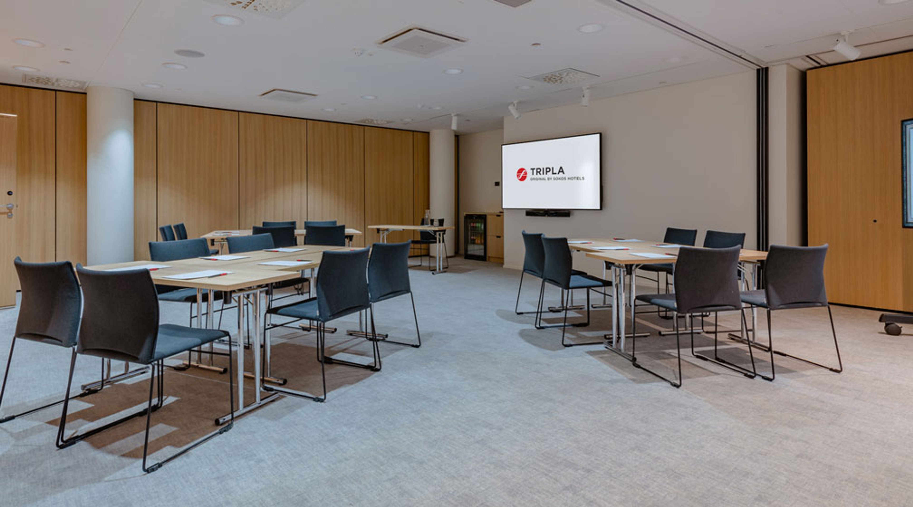 Moderni kokoustila puupaneeliseinineen, useita pöytiä ja tuoleja aseteltuna seminaarimuotoon, valkokangas edessä, tasainen lattiapinta.