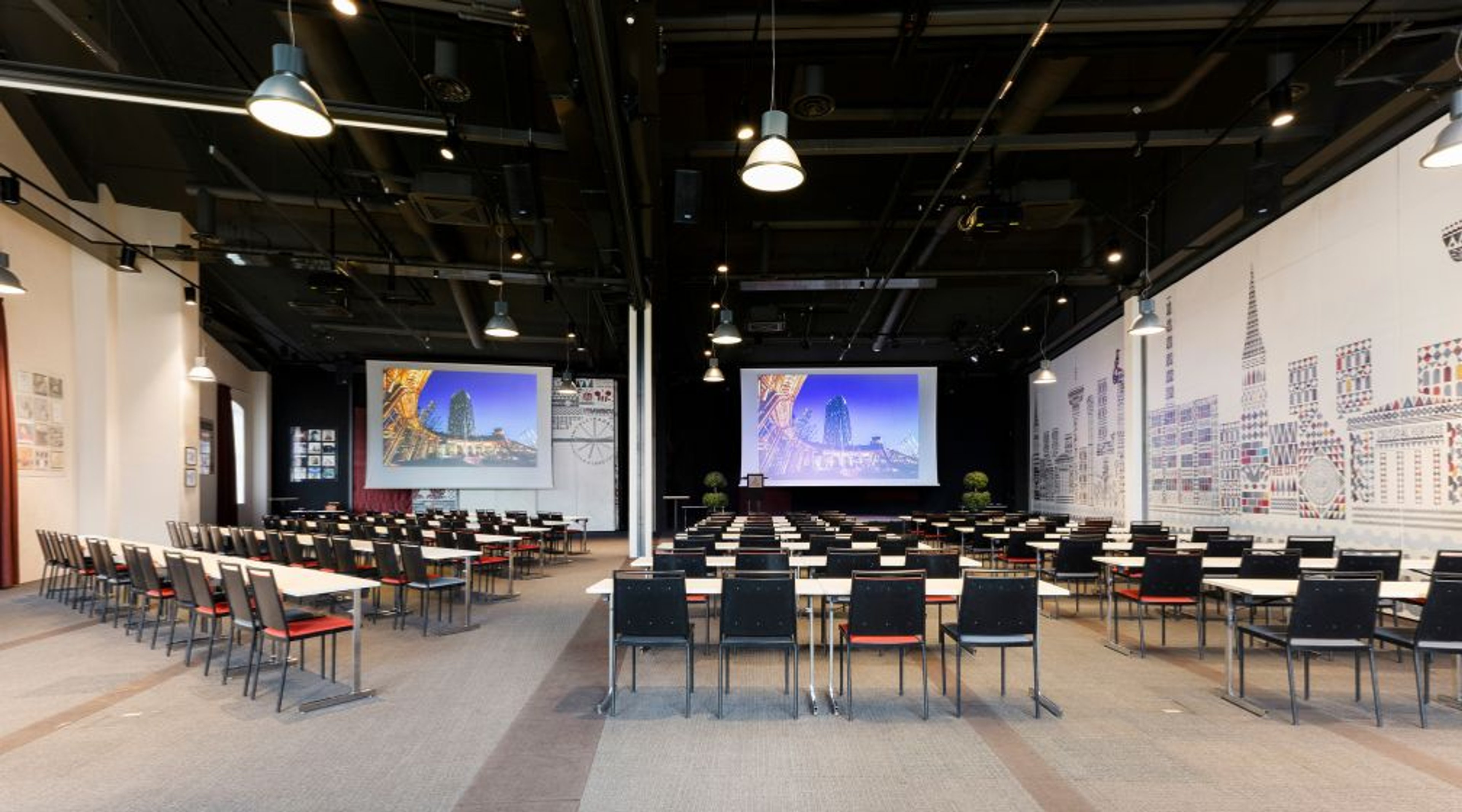 Konferenssitila, jossa rivit pöytiä ja tuoleja, kaksi suurta näyttöä, musta kattopinta ja riippuvalaisimet.