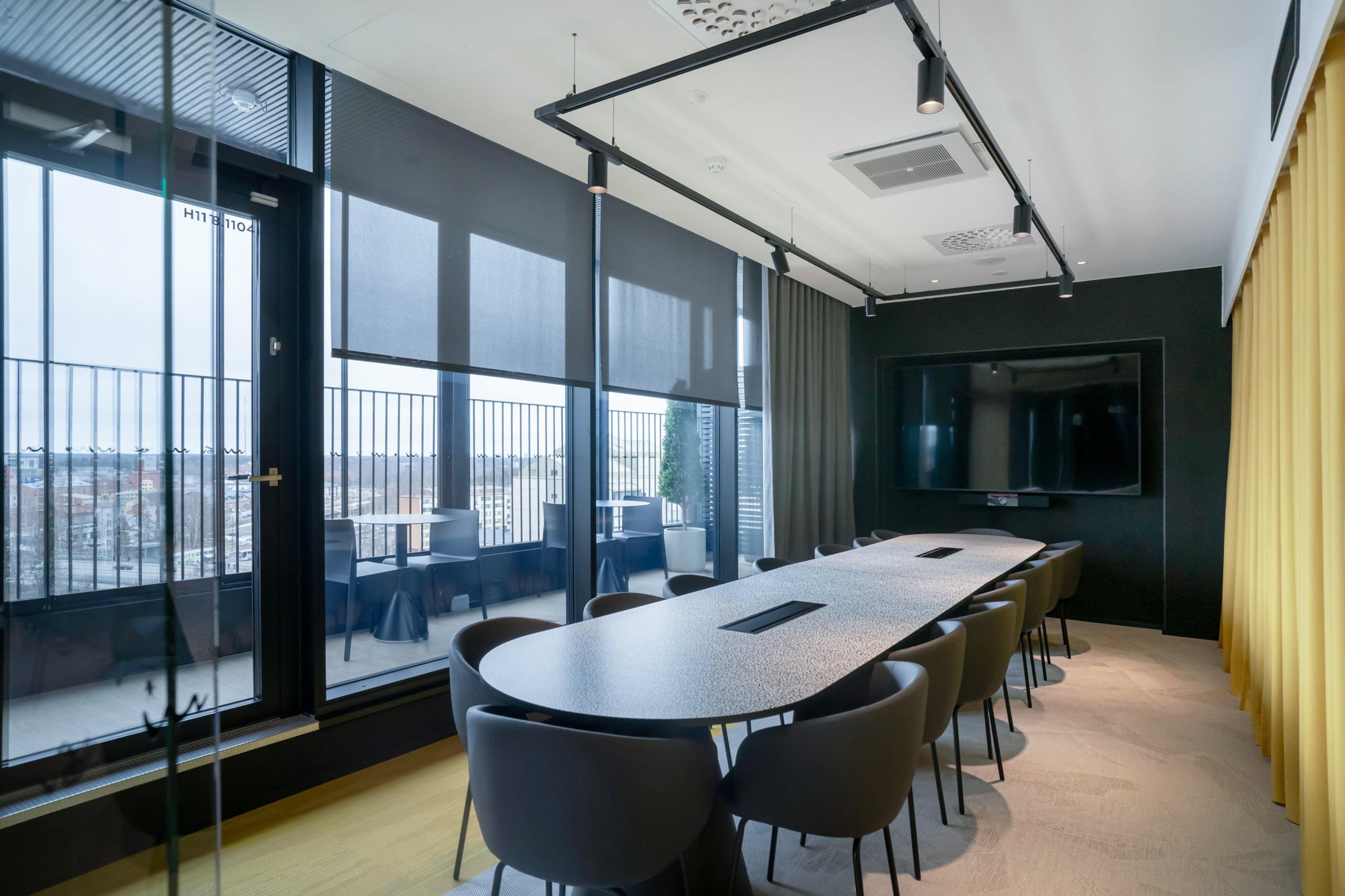 Moderni neuvotteluhuone, jossa pitkä pöytä, harmaat tuolit, suuri näyttö seinällä ja lattiasta kattoon ulottuvat ikkunat kaupunkinäkymällä.