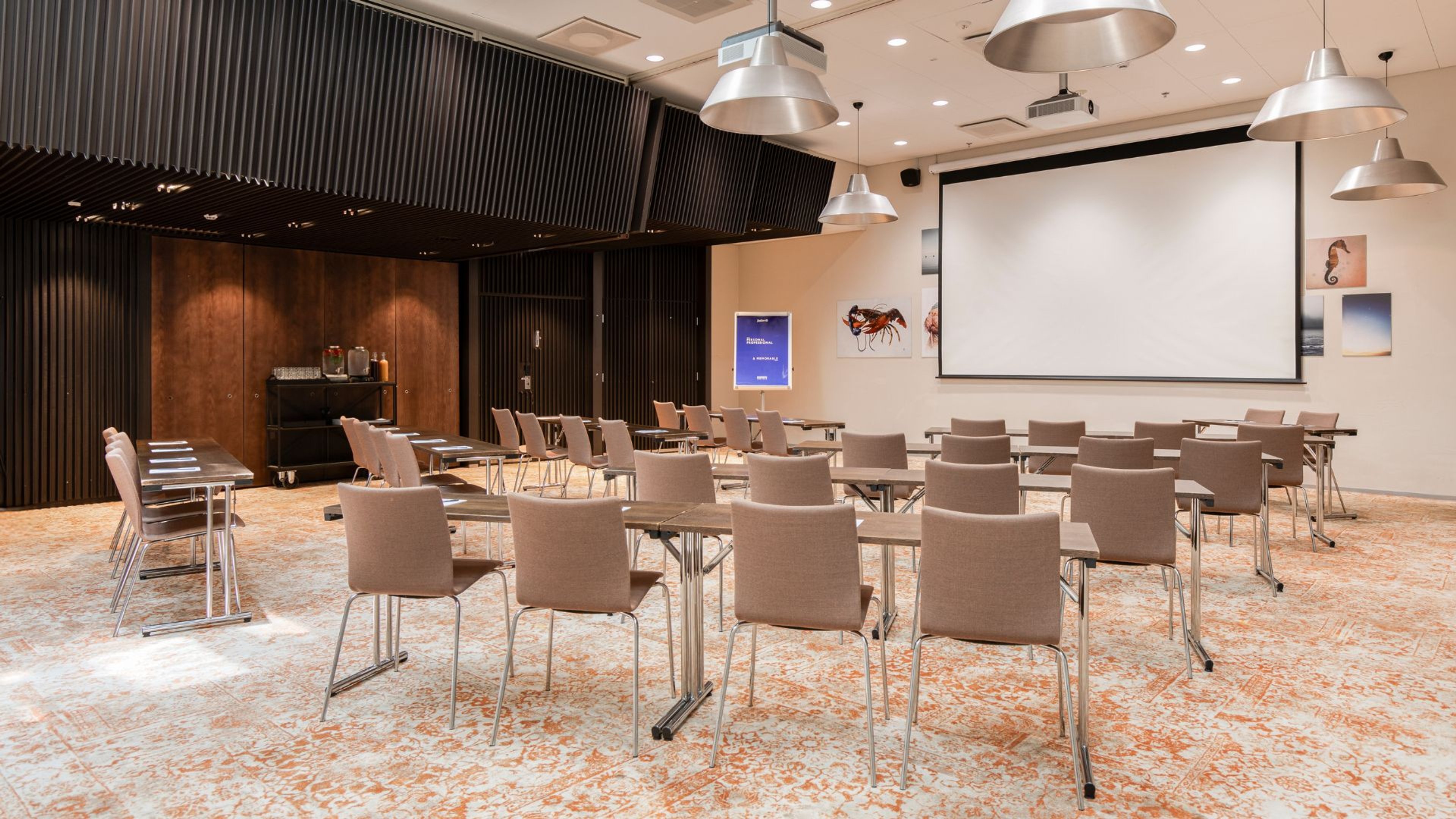 Moderni kokoustila pöytäryhmillä, projektiokankaalla ja tummalla puupaneloinnilla. Tasainen lattia ja hyvin valaistu tila.