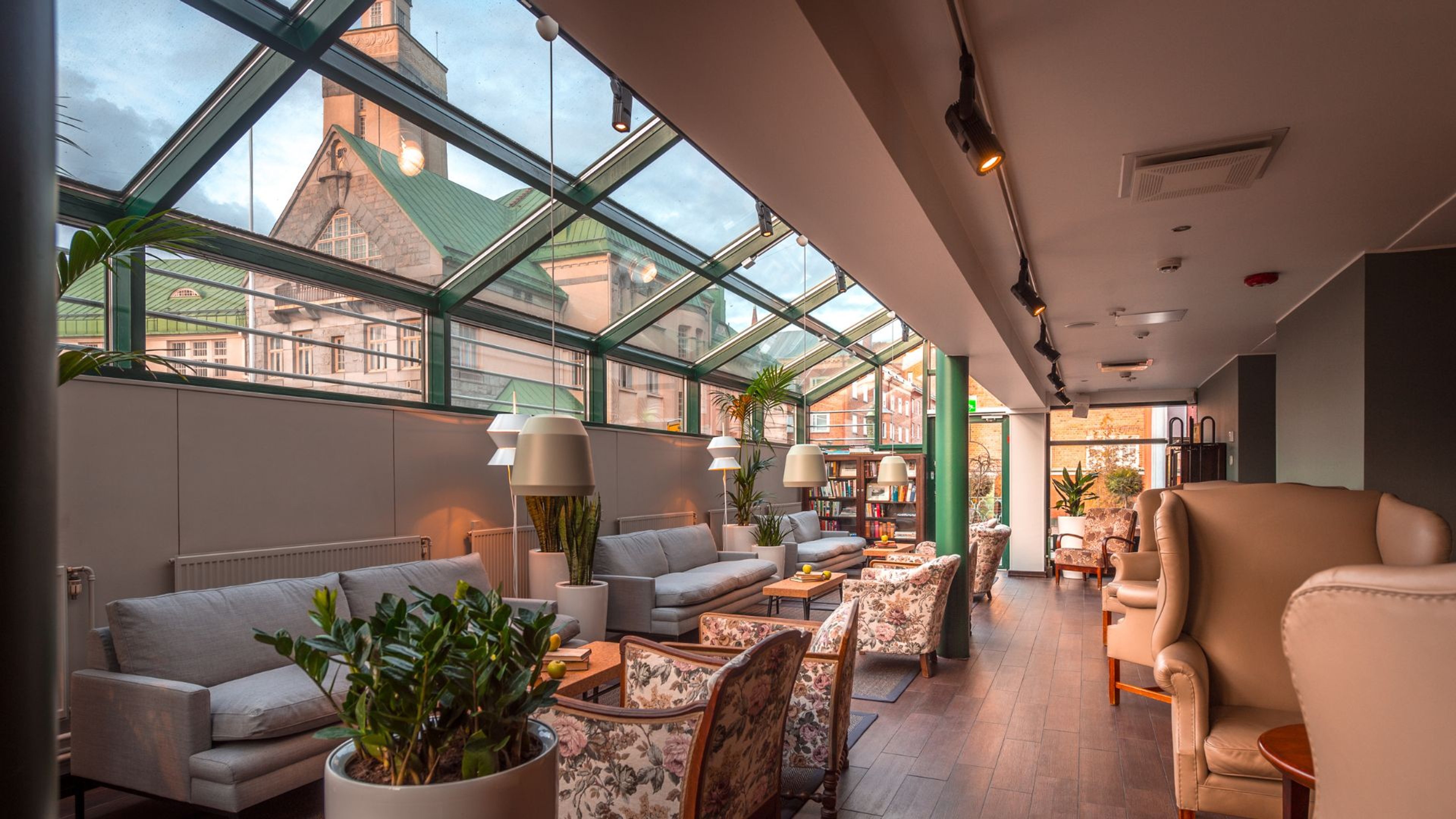 Valoisa oleskelutila lasiseinäisellä käytävällä. Sisustettu sohvilla, nojatuoleilla ja viherkasveilla. Puulattia ja näkymä historialliseen rakennukseen.