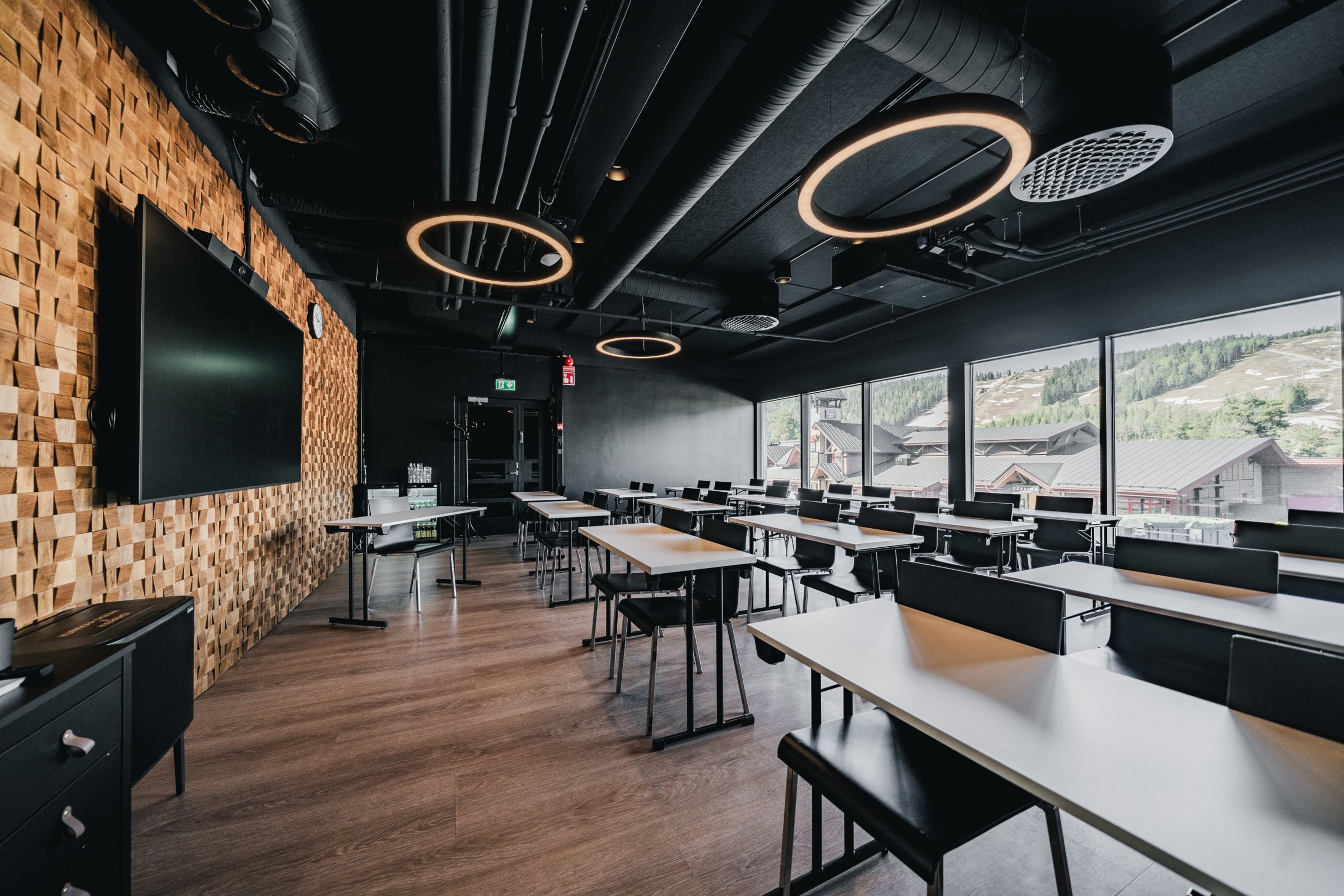 Moderni kokoustila puuseinällä, valkoisia pöytiä, mustia tuoleja ja suuret ikkunat vuoristomaisemalla. Puulattia ja pyöreät kattovalaisimet.