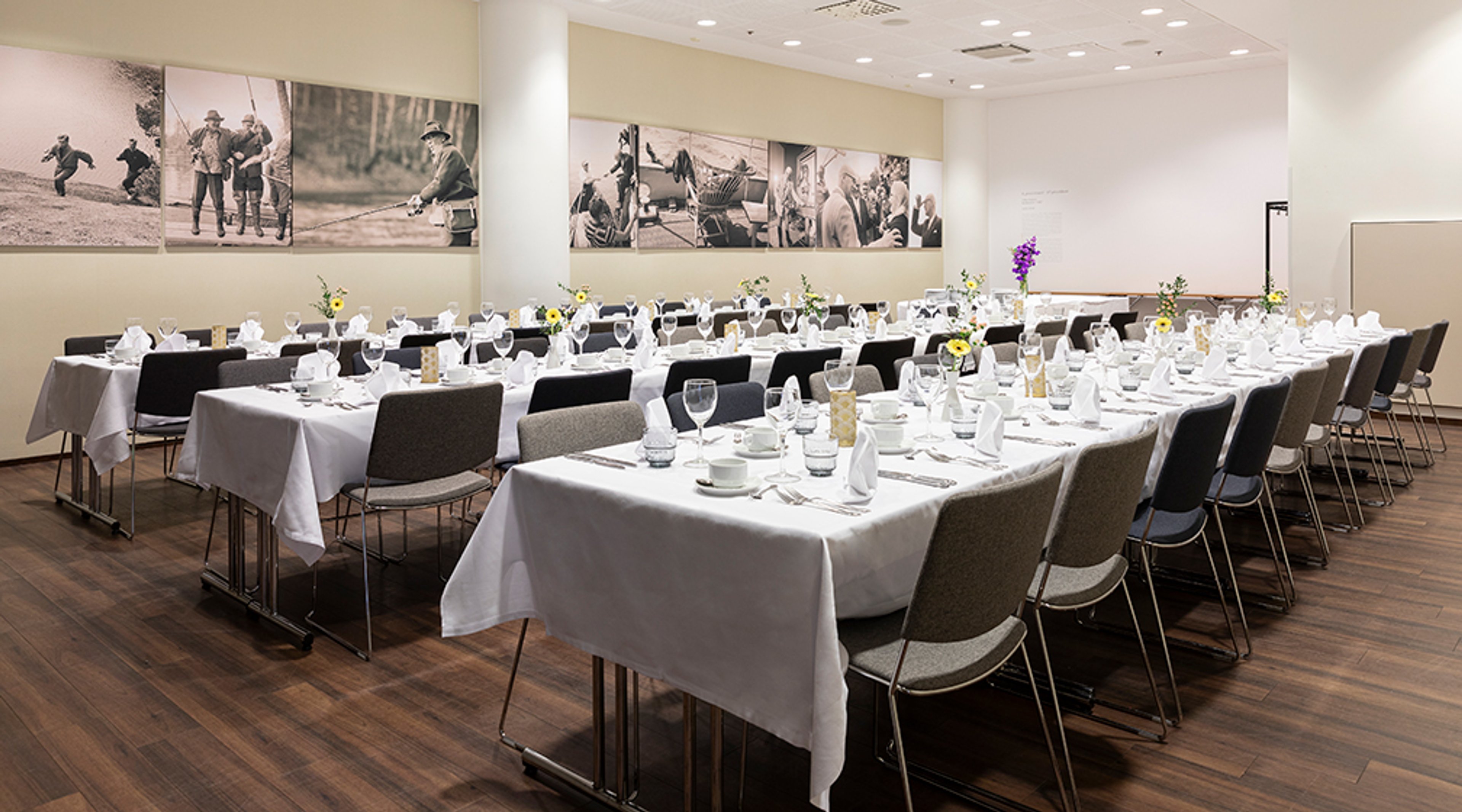 Juhlatila pitkillä pöydillä, valkoisilla liinoilla ja kattauksilla. Vaaleilla seinillä mustavalkoisia valokuvia. Tilassa puulattia ja harmaat tuolit.