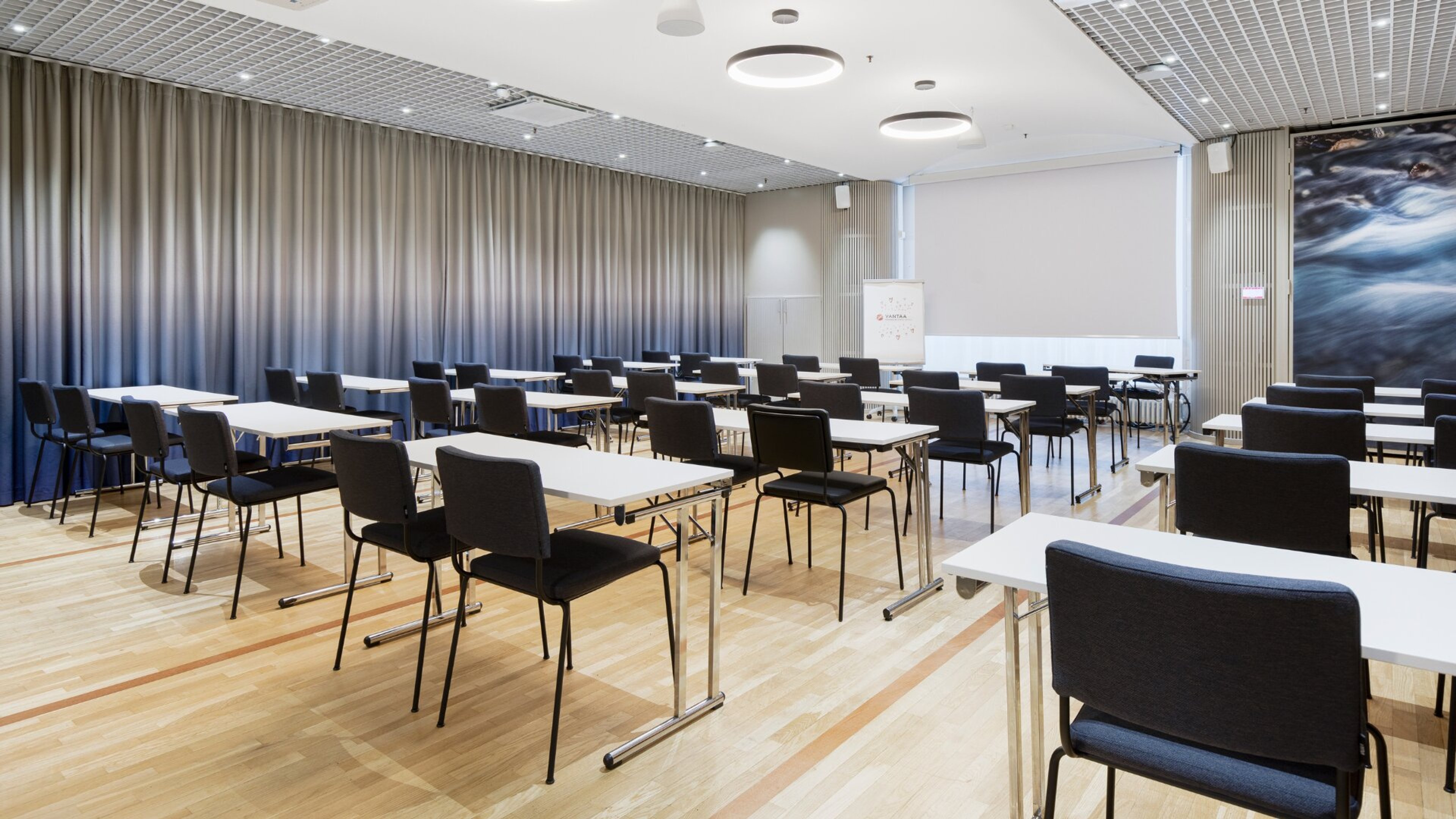 Moderni kokoustila valkoisilla pöydillä ja mustilla tuoleilla. Tila on avara, hyvin valaistu ja lattialla on parketti.