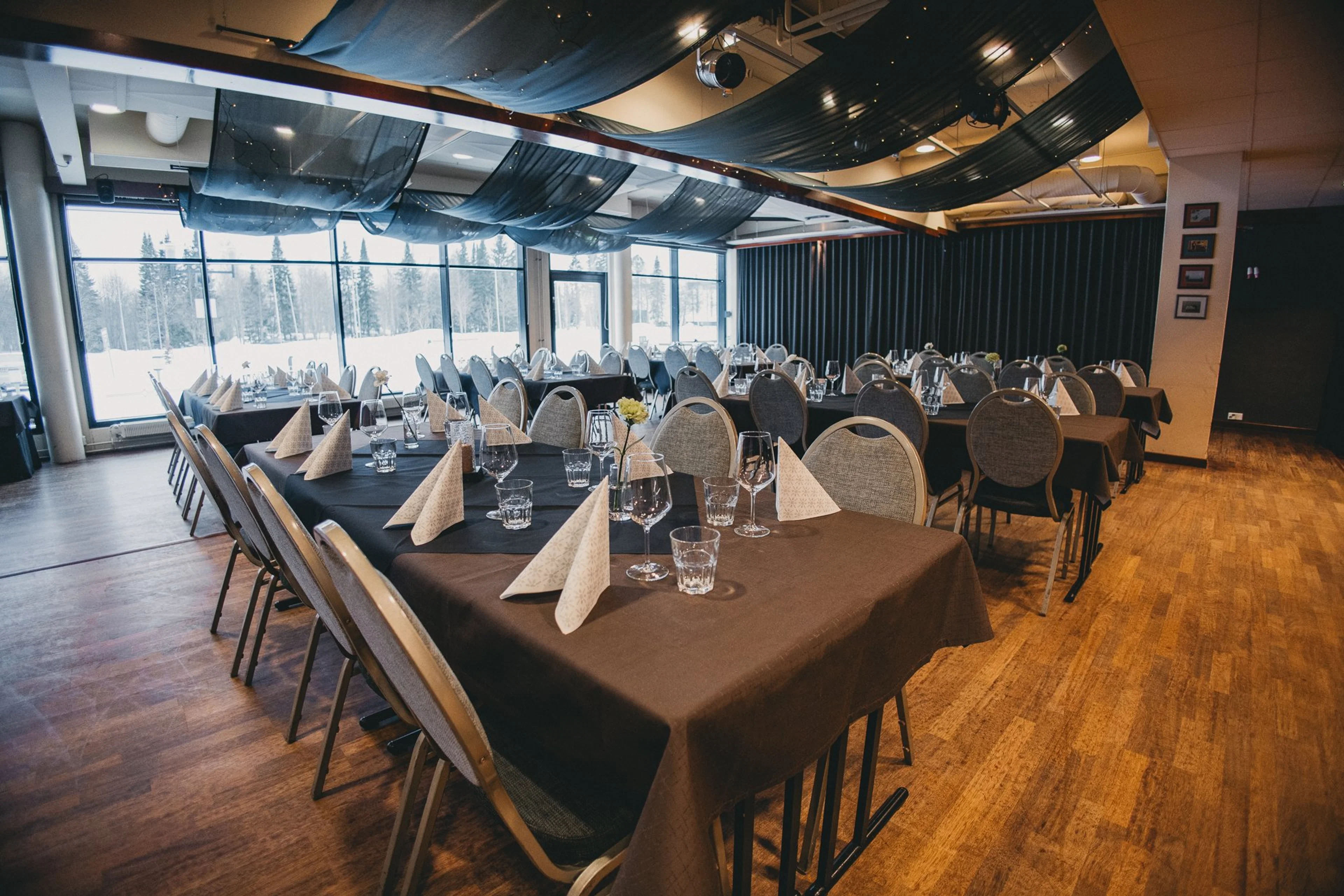 Juhlatila, jossa pöytiä mustilla pöytäliinoilla ja taitetut servietit. Lattialla puuparketti. Isot ikkunat näyttävät lumiseen maisemaan.