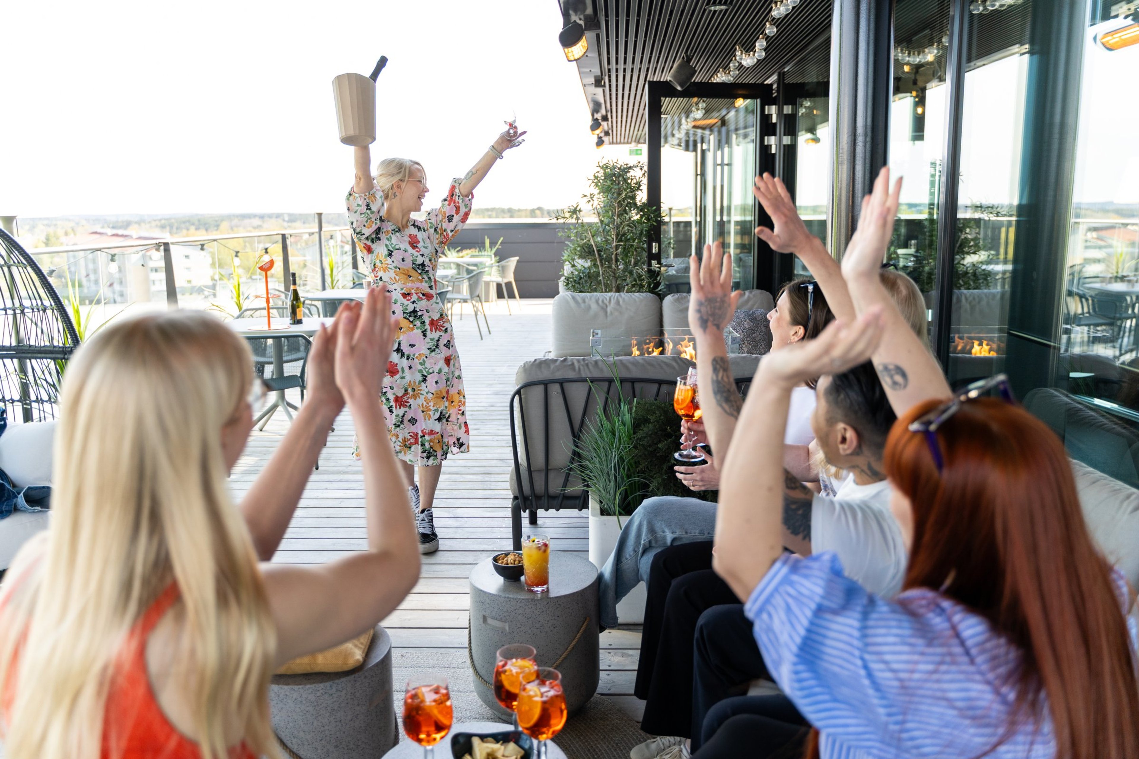 Henkilöt juhlimassa kattoterassilla, jossa puinen lattia ja lasikaiteet. Taustalla näkyy kaupunkimaisema.