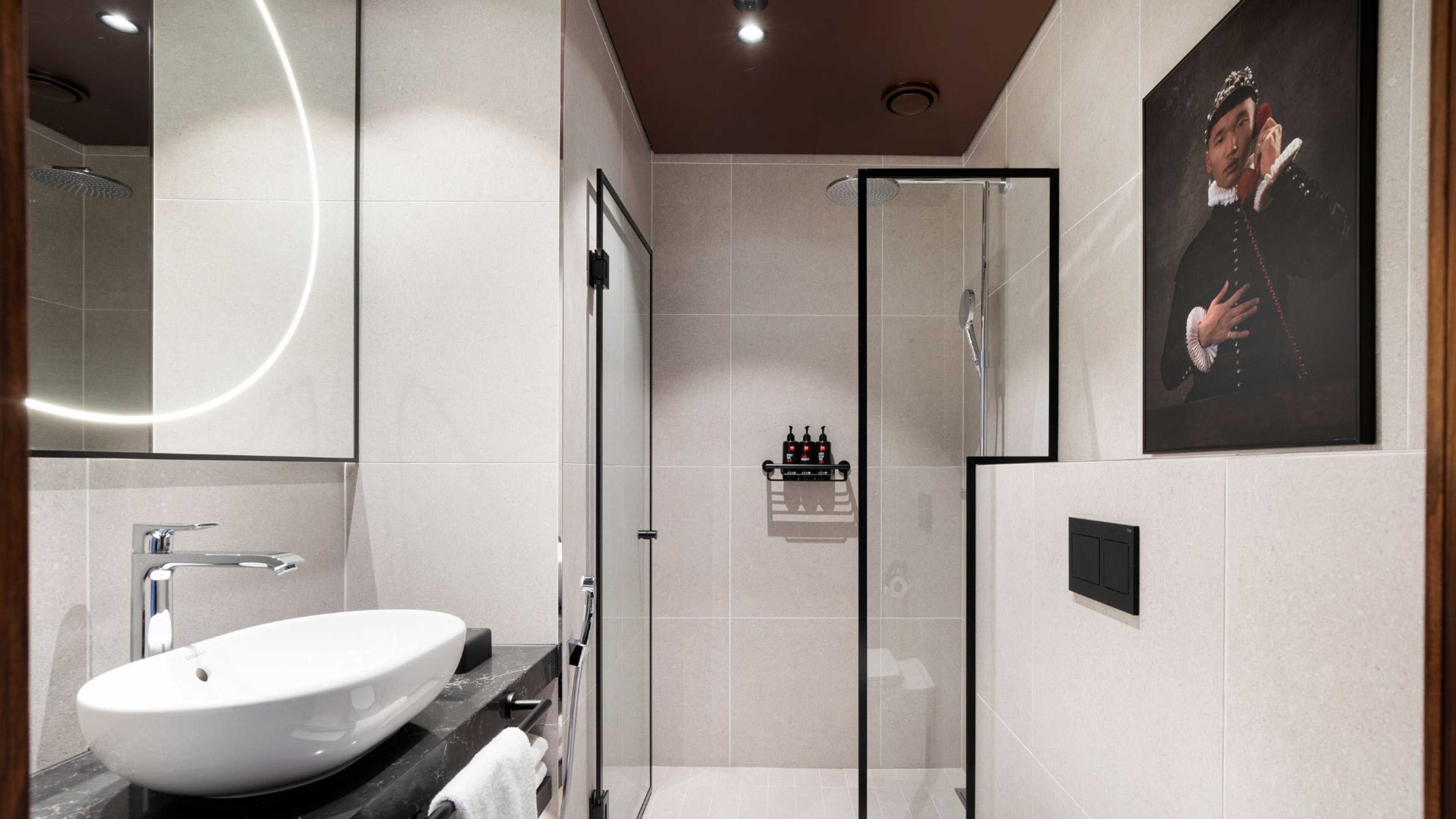 Moderni kylpyhuone, jossa on valkoinen pesuallas, tasopeili valaistuksella, lasiseinäinen suihku ja historiallinen muotokuva seinällä.