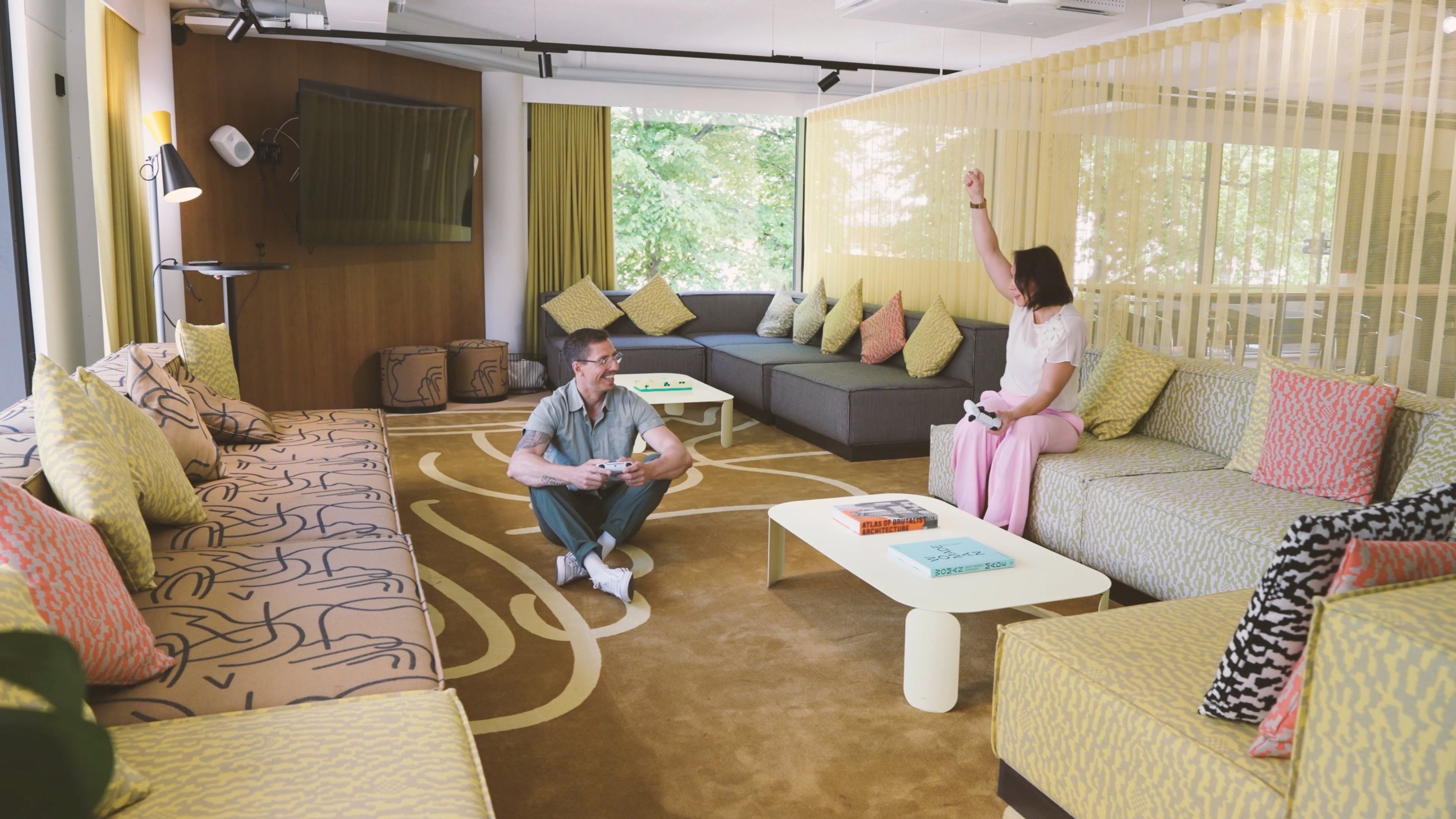 Moderni oleskelutila useilla sohvilla ja värikkäillä tyynyillä. Tilava huone jossa isot ikkunat, patternikuviollinen matto ja kaksi henkilöä pelaavat videopelejä.