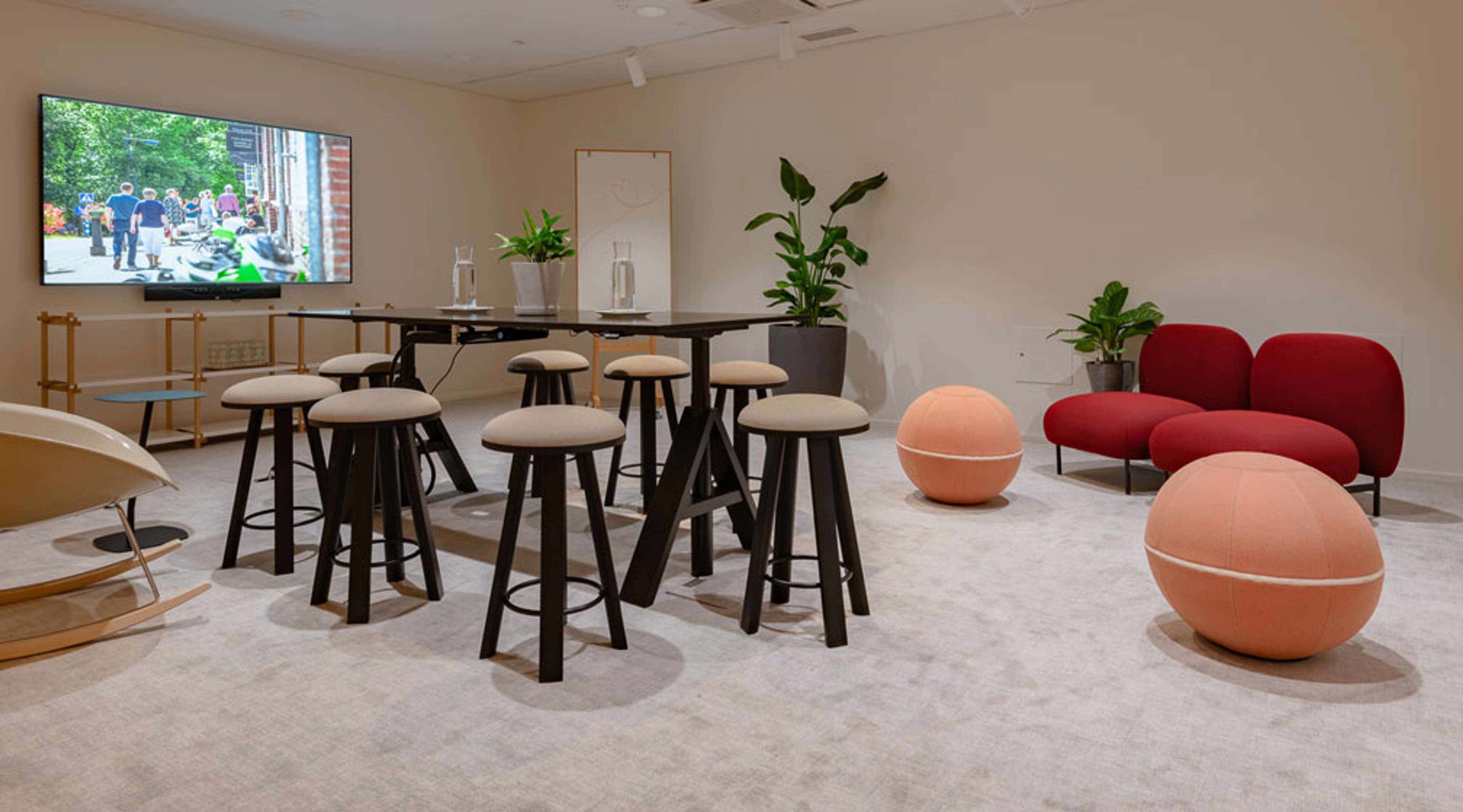 Moderni neuvottelutila, jossa baaripöytä jakkaroilla, punainen sohva, vaaleanpunaiset istuinpallot, viherkasvia ja suuri näyttö seinällä. Vaaleat matot lattialla.