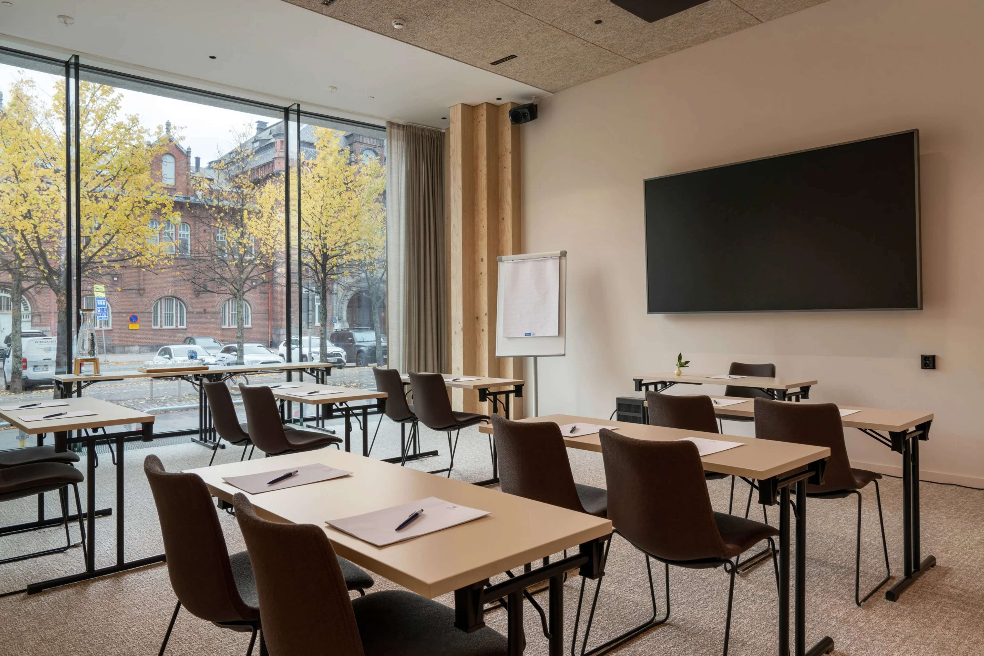 Modernt mötesrum med bord och stolar, stor skärm på väggen och golv-till-tak-fönster med utsikt över gula träd på gatan.
