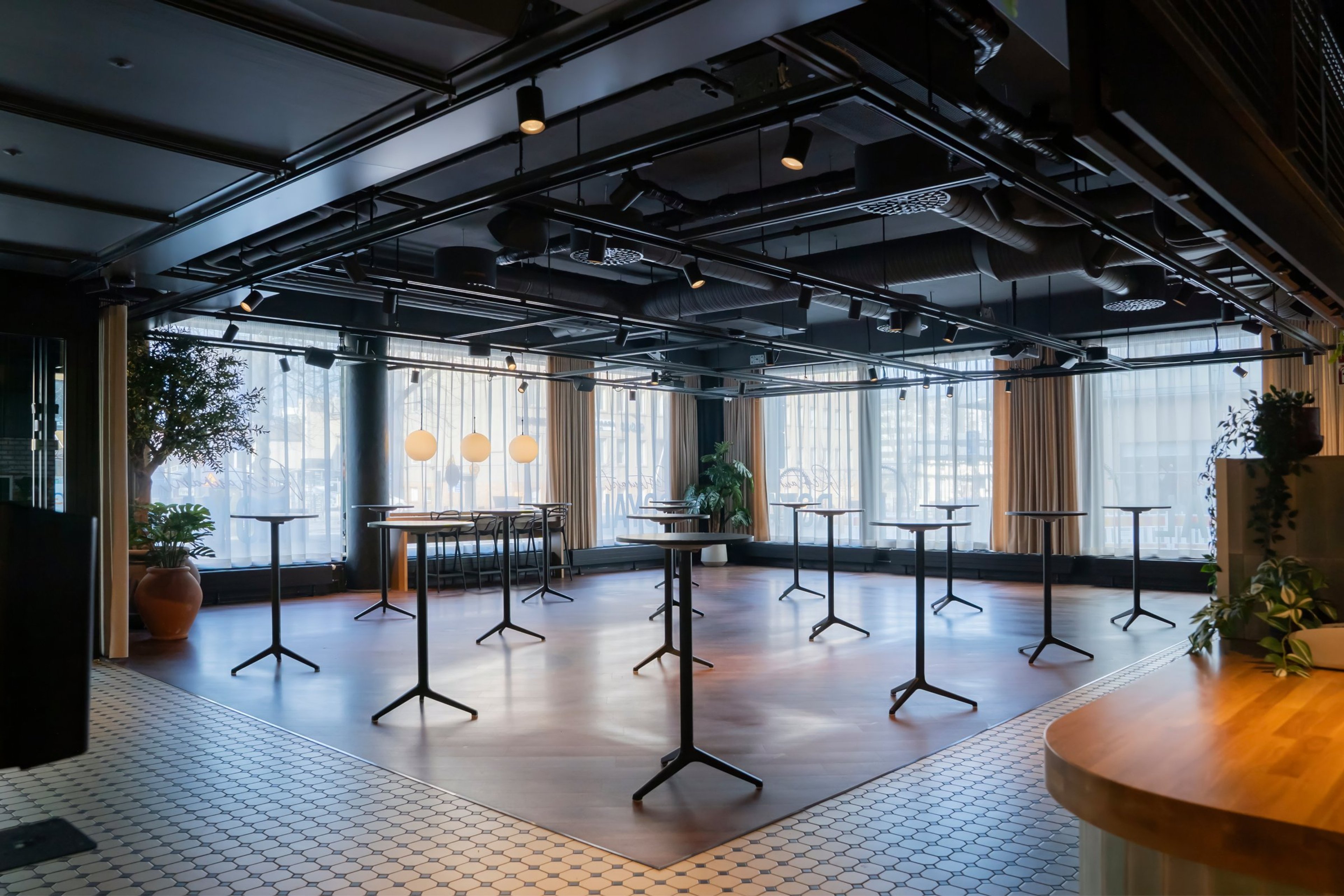 Moderni tapahtumatila, jossa korkeita pöytiä avoimella lattialla. Tilassa isot ikkunat, beige verhot, kasveja nurkissa ja tasainen puulattia.