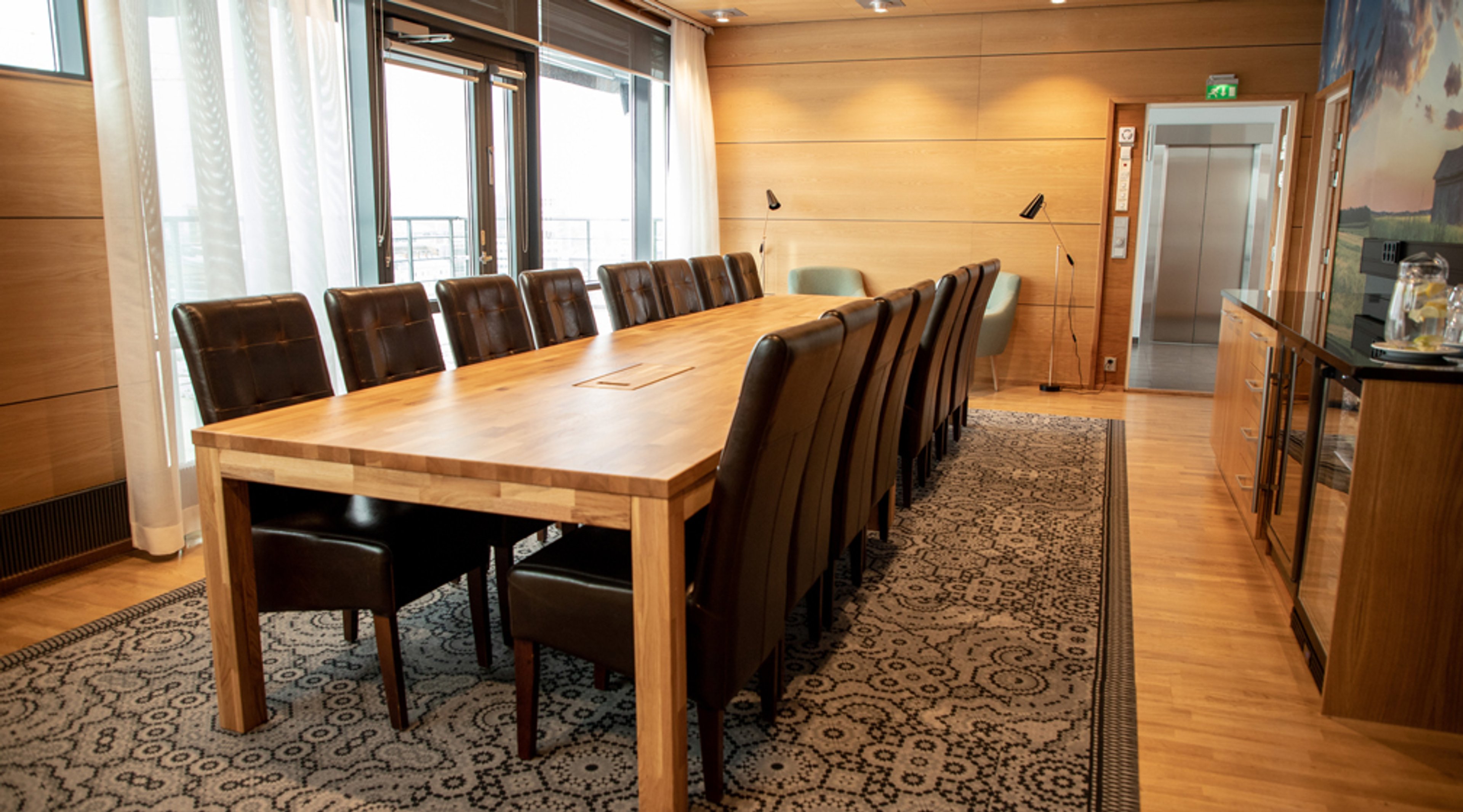 Kokoushuone, jossa pitkä puinen pöytä, nahkatuoleja, puulattiaa, ikkunat, koristeellinen matto ja hissi takaosassa.