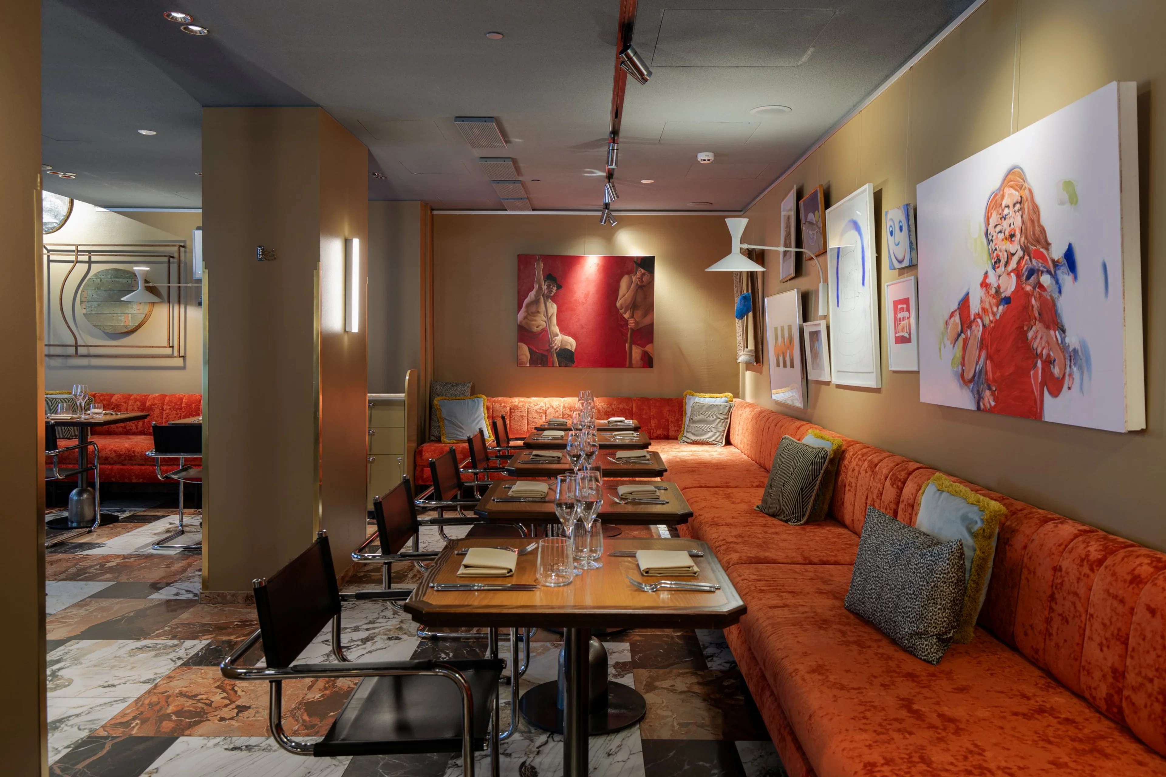 Tyylikkään ravintolan ruokailusali, jossa oranssi sohva seinän vierellä, pöytiä katettuna ja taideteoksia seinillä. Marmorilattia ja hyvä valaistus.