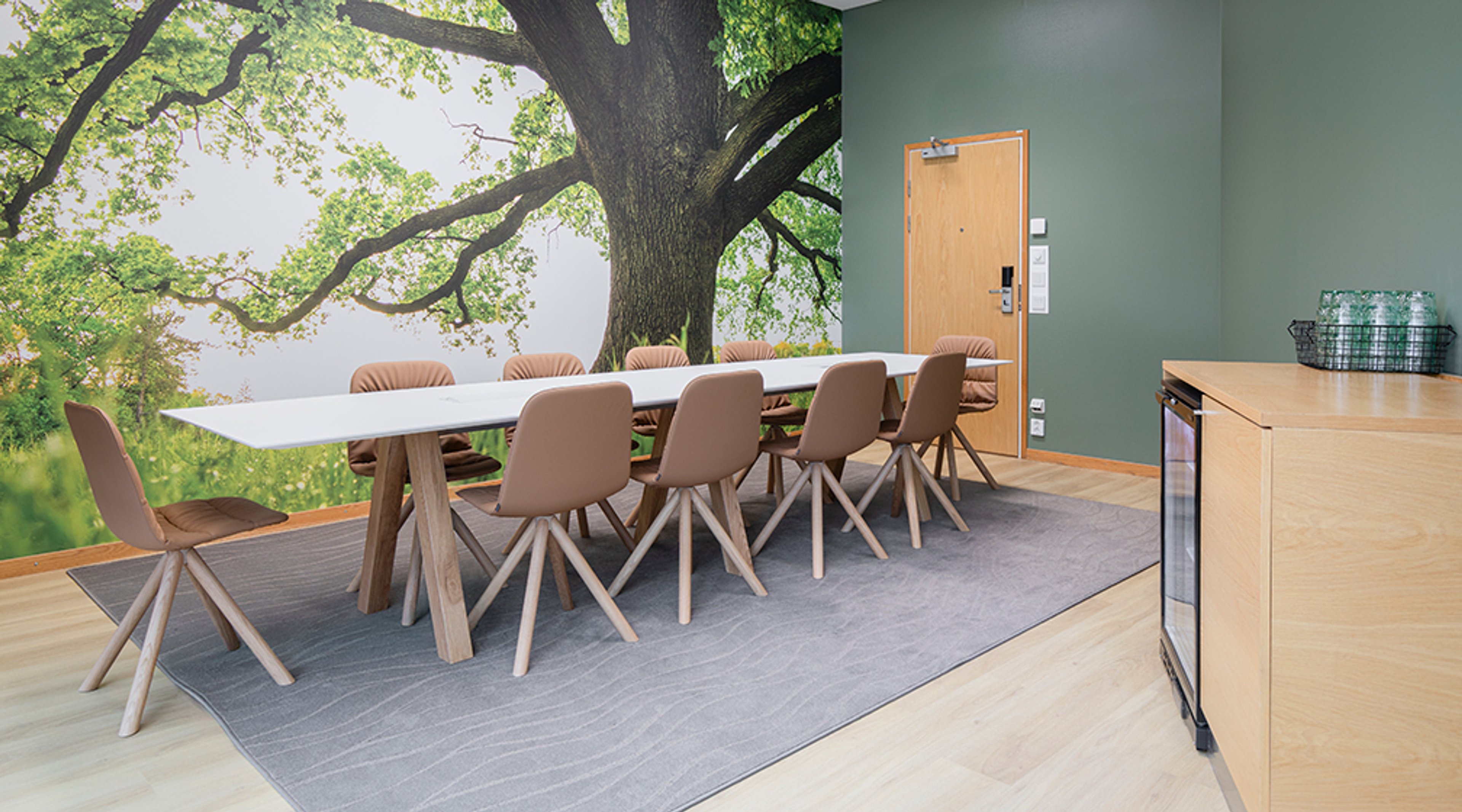 Neuvotteluhuone, jossa pitkä pöytä, ruskeita tuoleja ja vihreällä seinällä puutapetti. Tilassa on puulattia, harmaa matto ja puinen kaappi.