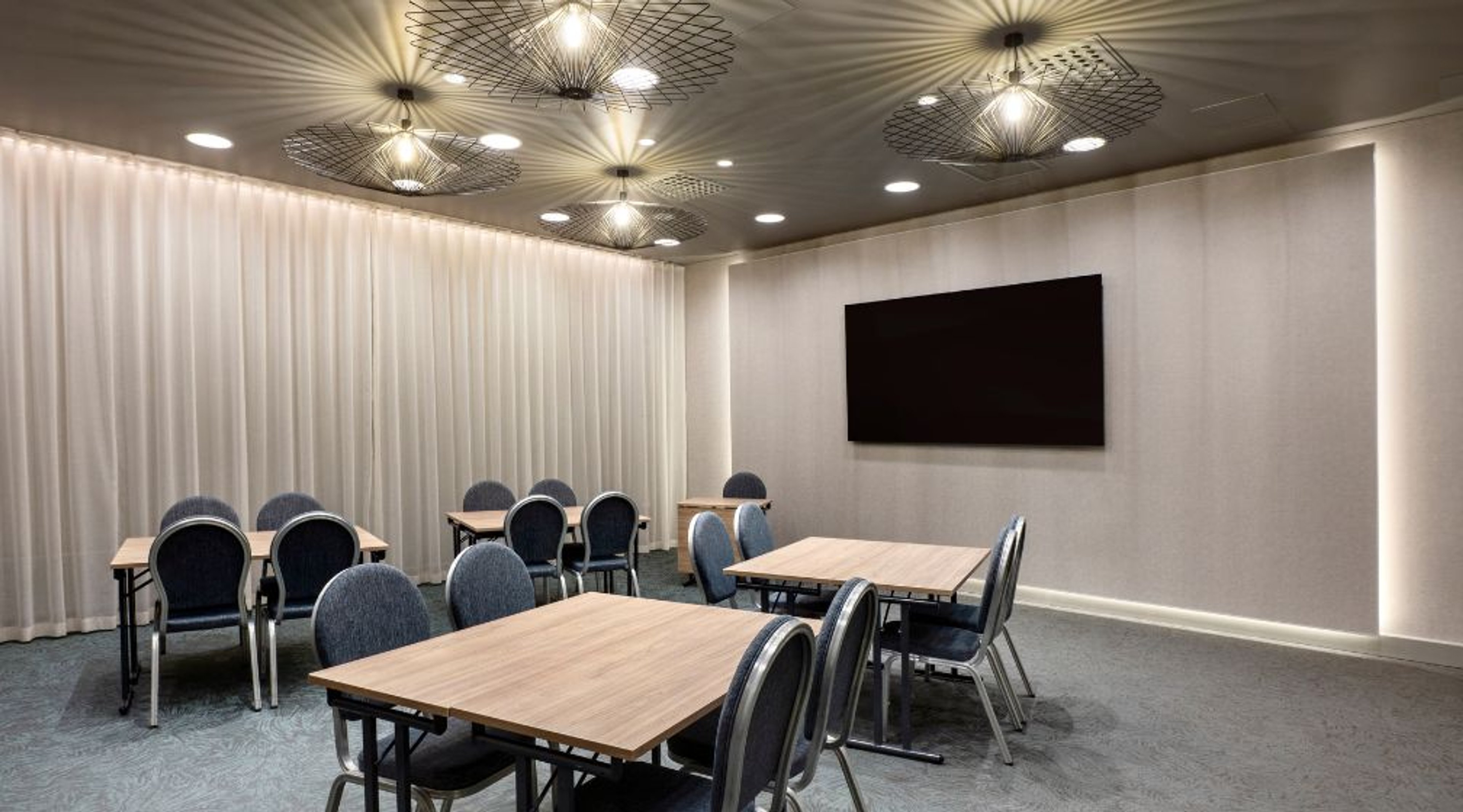 Moderni neuvotteluhuone, jossa kolme puupöytää, harmaalla verhoillut tuolit, vaalea sisustus, tyylikkäät kattovalaisimet ja näyttö seinällä.