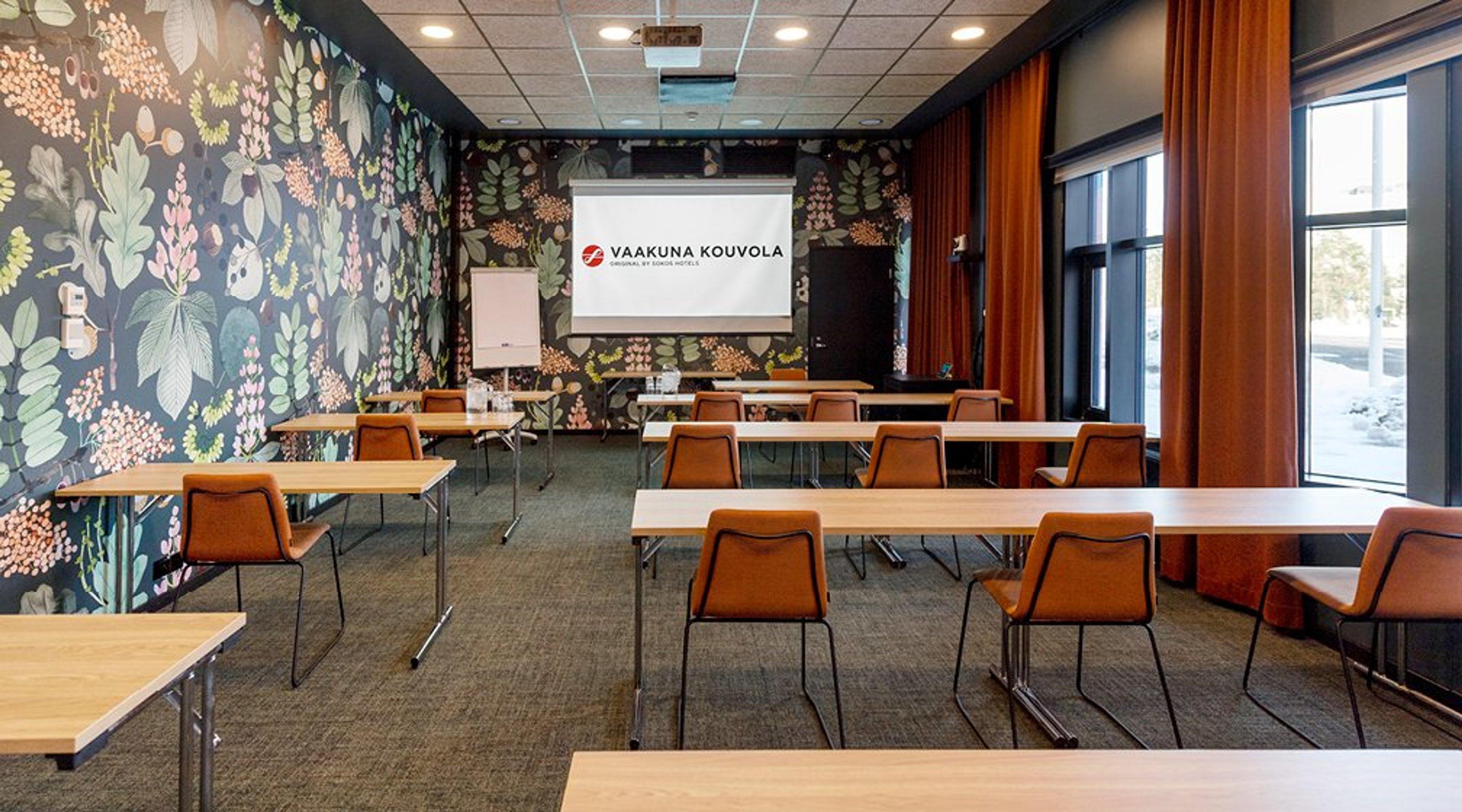 Kokoustila, jossa puiset pöydät ja oranssit tuolit. Seinillä värikkäät kukkatapetit, oranssit verhot ja edessä valkea projektorikangas.