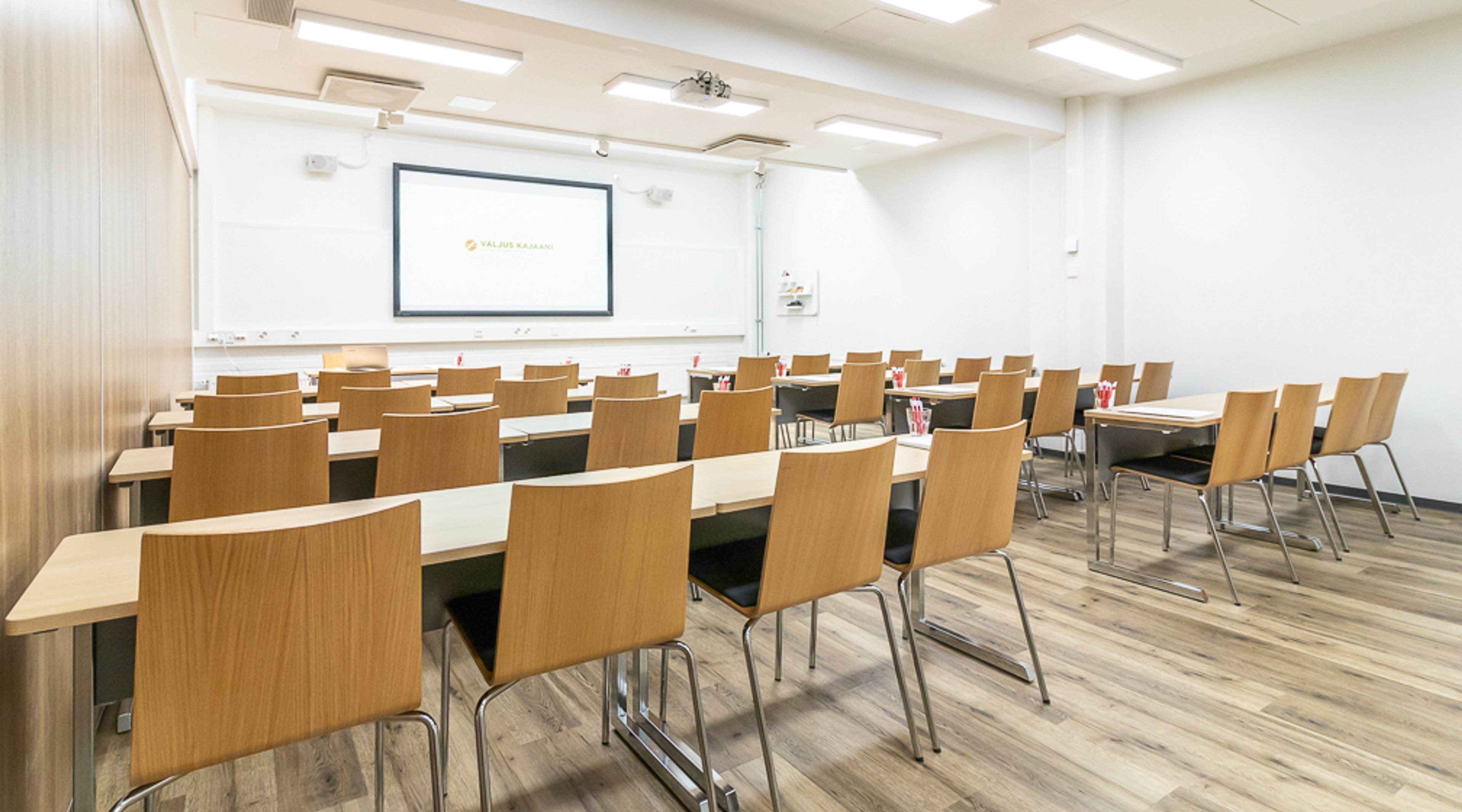 Luokkahuonemainen kokoustila puisilla tuoleilla, valkoisilla pöydillä ja projektorikankaalla edessä. Vaalea puulattia ja hyvä valaistus.
