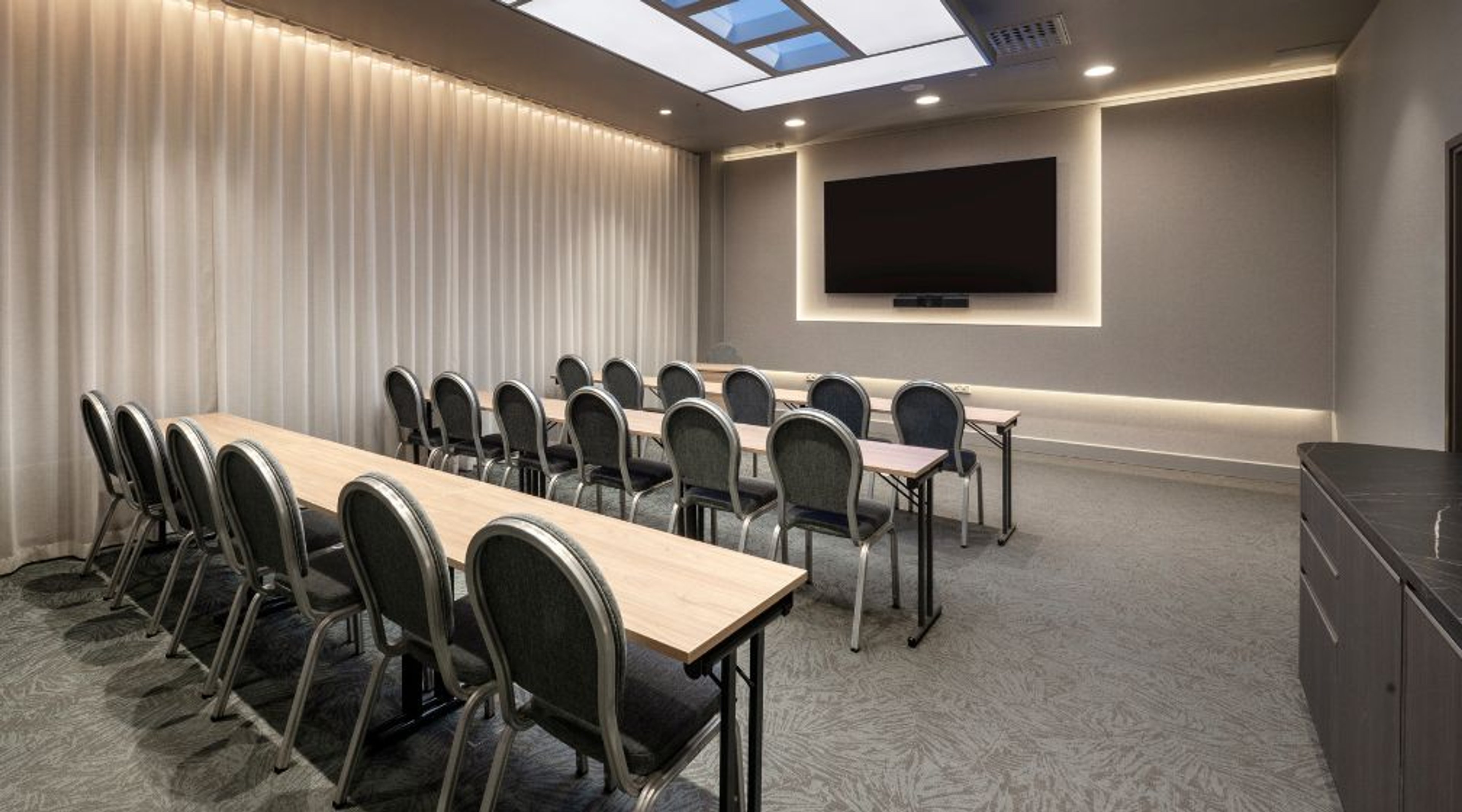 Moderni kokoushuone, jossa pitkät pöydät, tuolit, suuri näyttö seinällä ja kattoikkuna. Tasainen harmaa lattiapinta ja valaistut seinät.