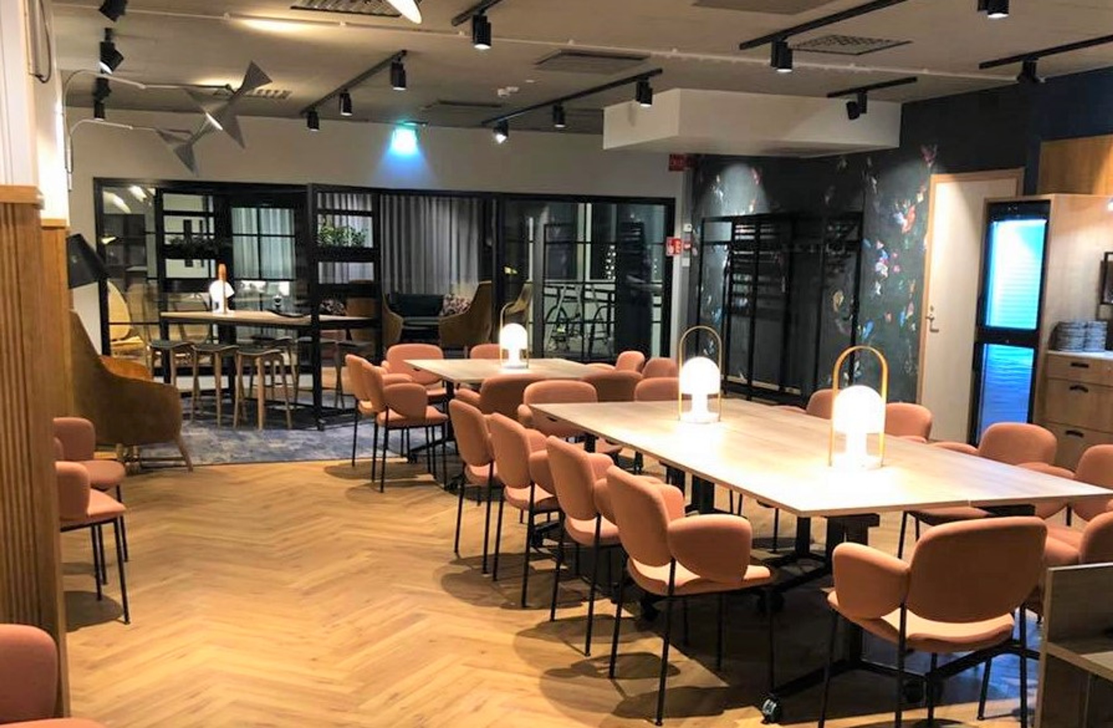 Moderni kokoustila vaaleanpunaisilla tuoleilla, puulattialla ja suurilla pöydillä. Avara tila, jossa lasiseiniä ja tyylikkäät valaisimet.