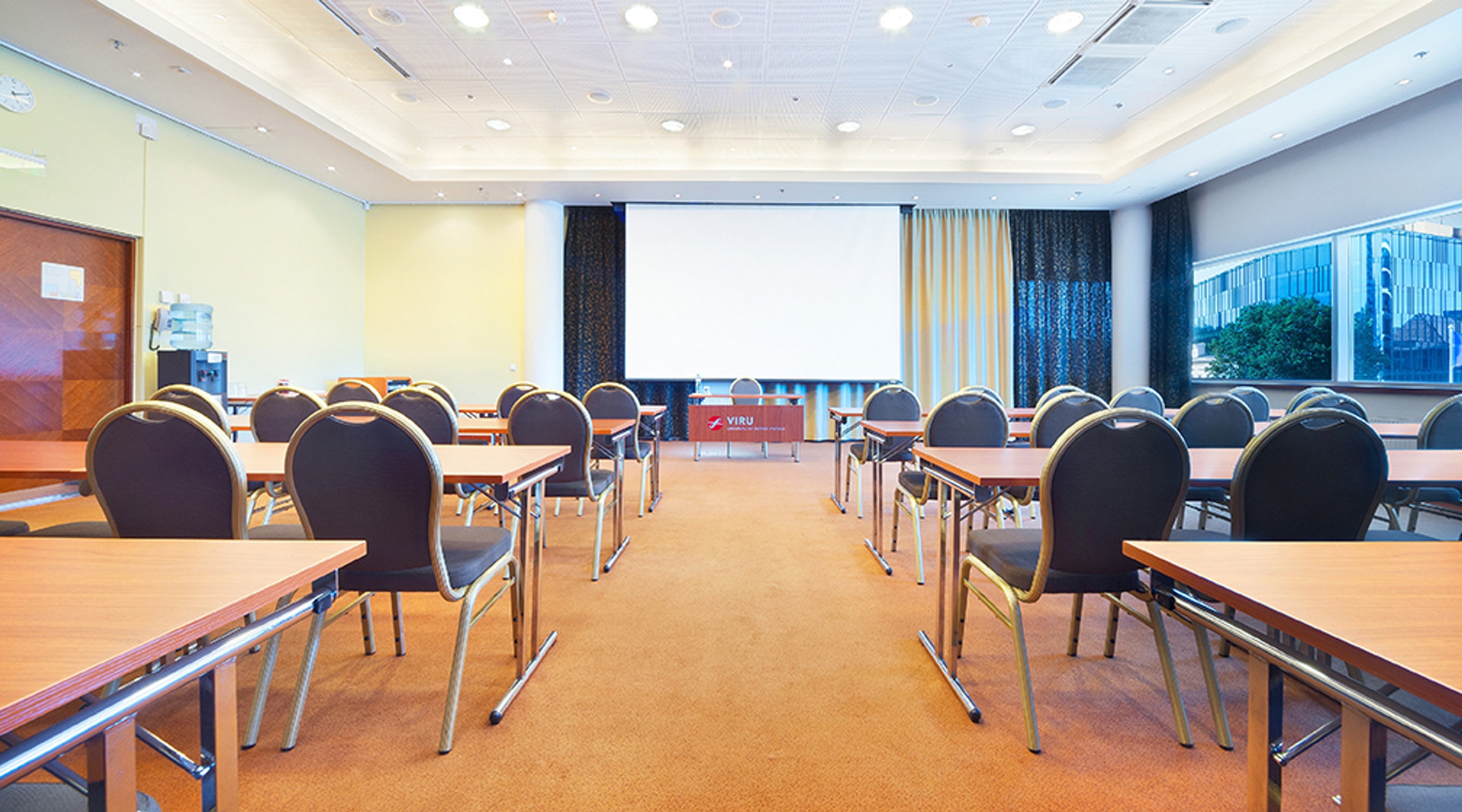 Kokoustila, jossa on useita puisia pöytiä ja mustia tuoleja. Huoneen edessä valkoinen projektorikangas ja taustalla ikkunanäkymä.