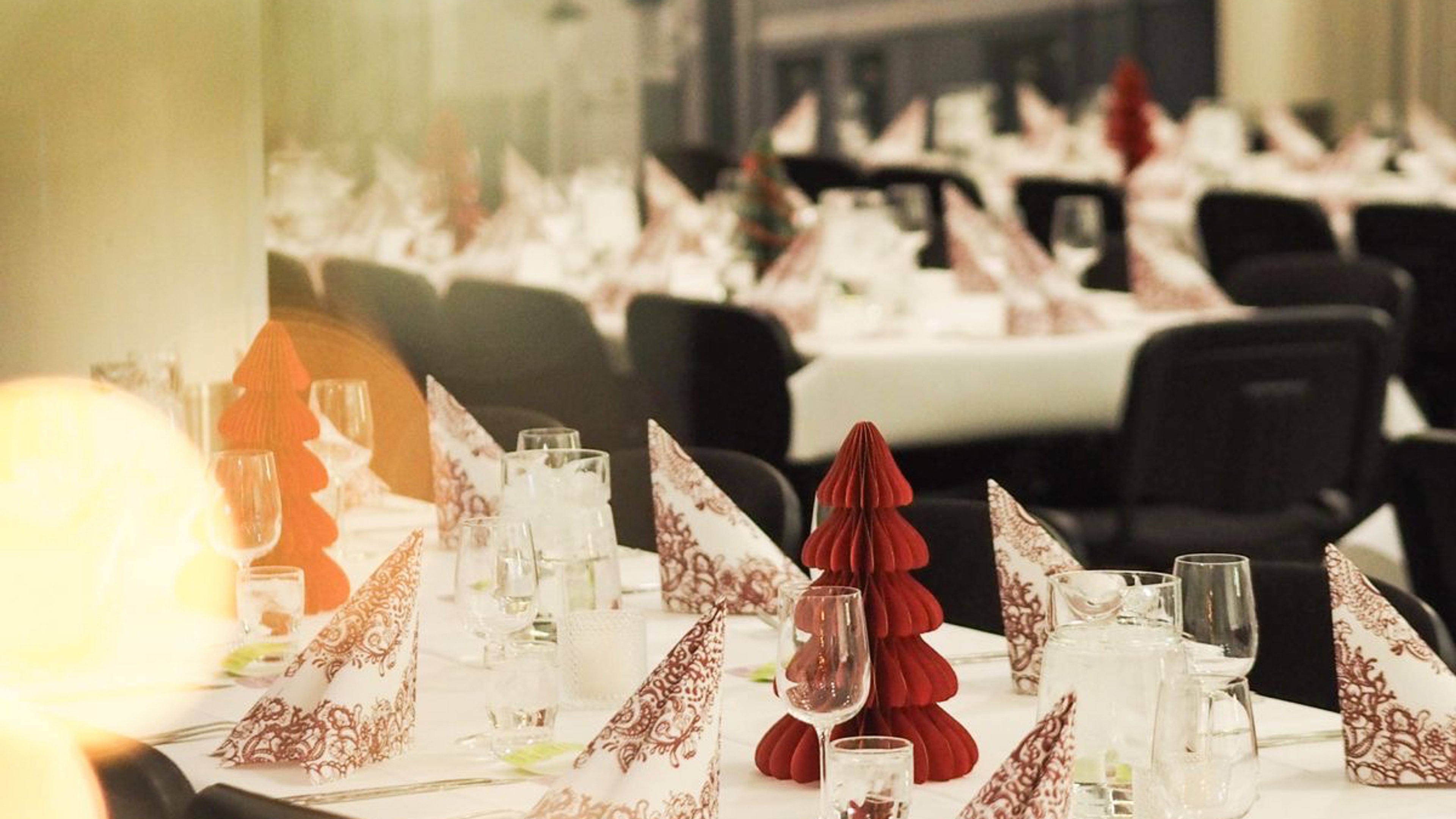 Restaurant tables decorated with red paper Christmas trees and ornate napkins. Glassware set for service.