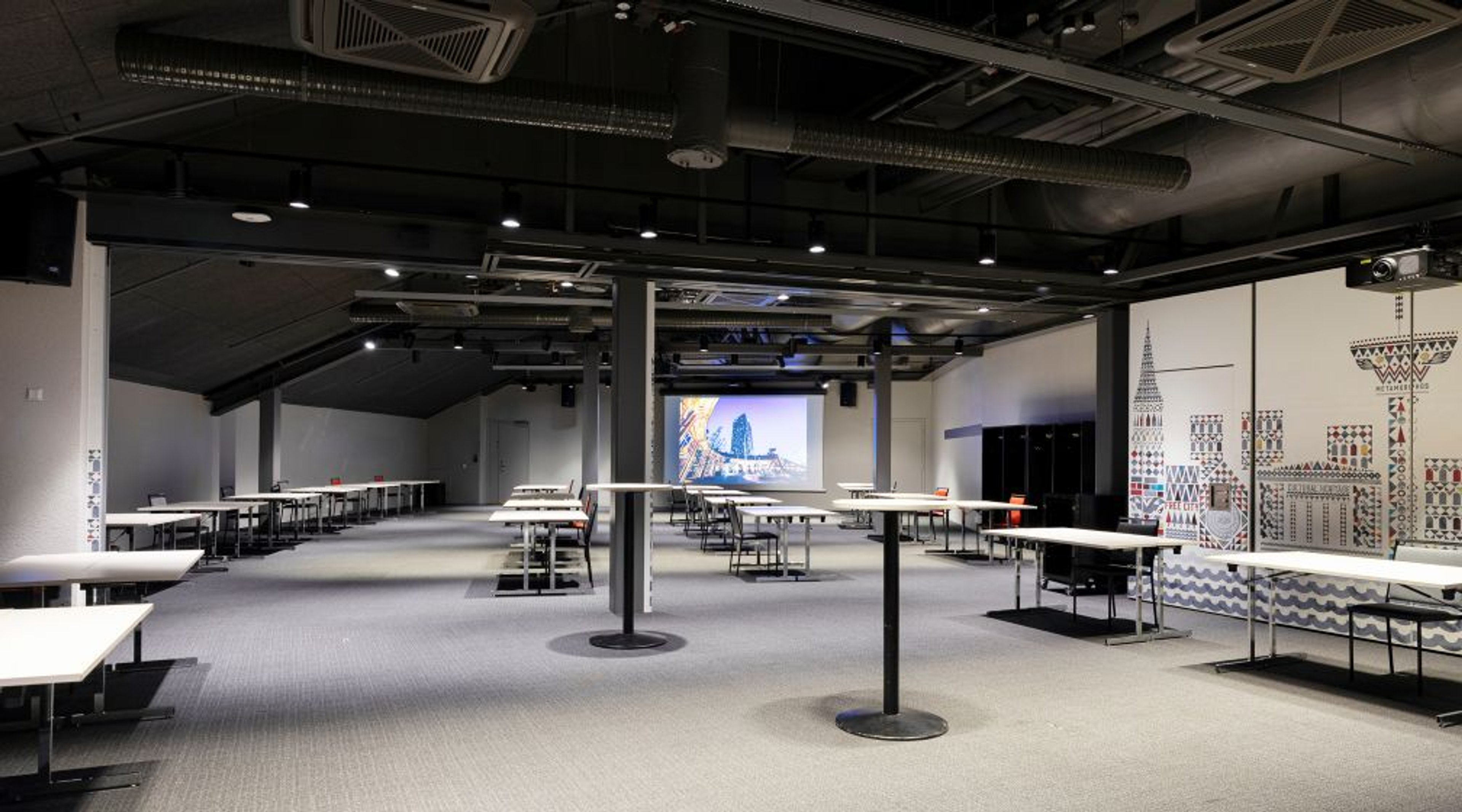 Avara konferenssi- tai koulutustila, jossa pöytiä ja tuoleja, projektorinäyttö takana, näkyvissä ilmanvaihtoputkisto katossa.
