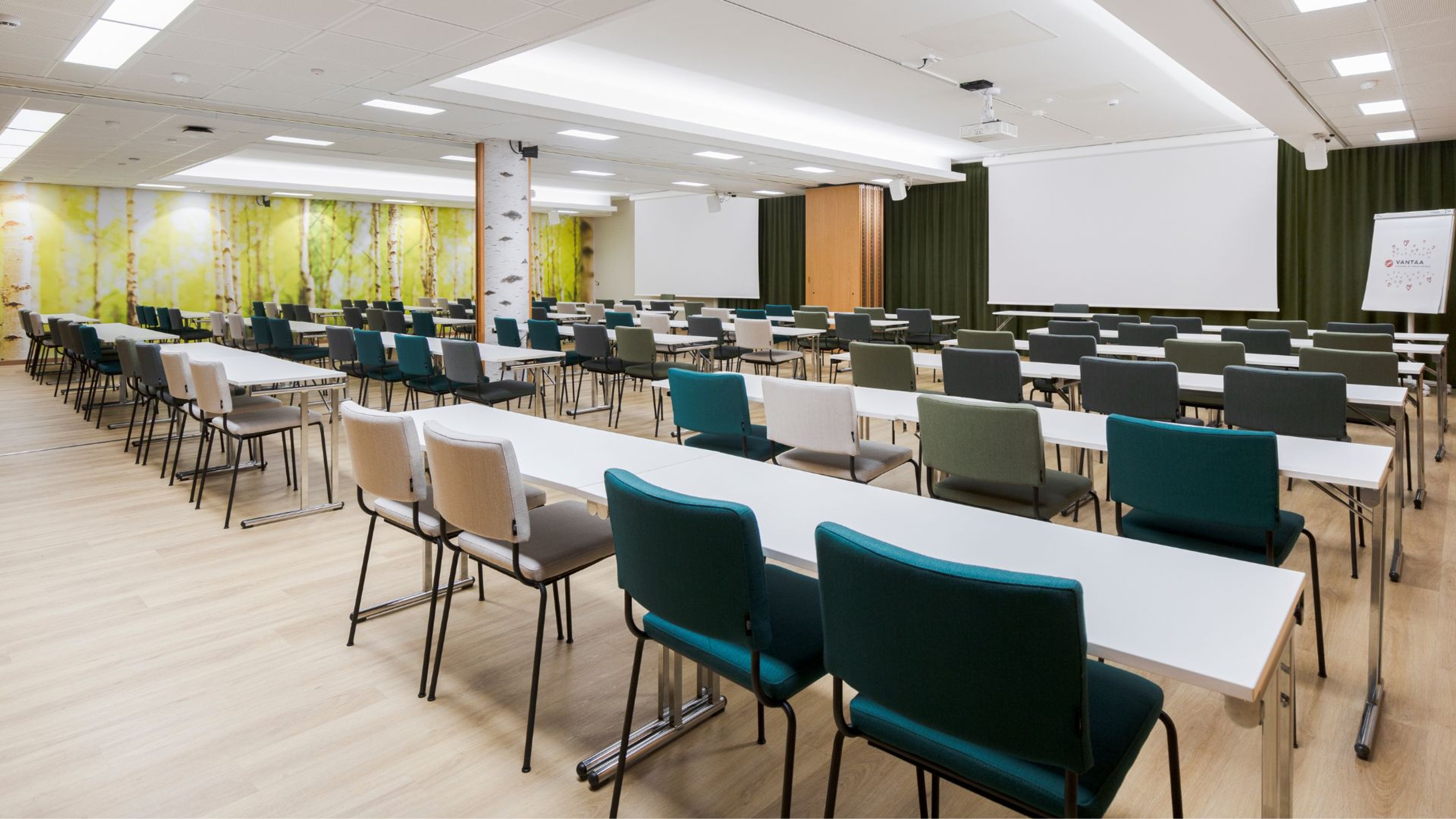 Valoisa kokoustila, jossa on pitkiä pöytiä, värikkäitä tuoleja, valkokankaita ja koivumetsätapetti. Tasainen kovapuulattia.