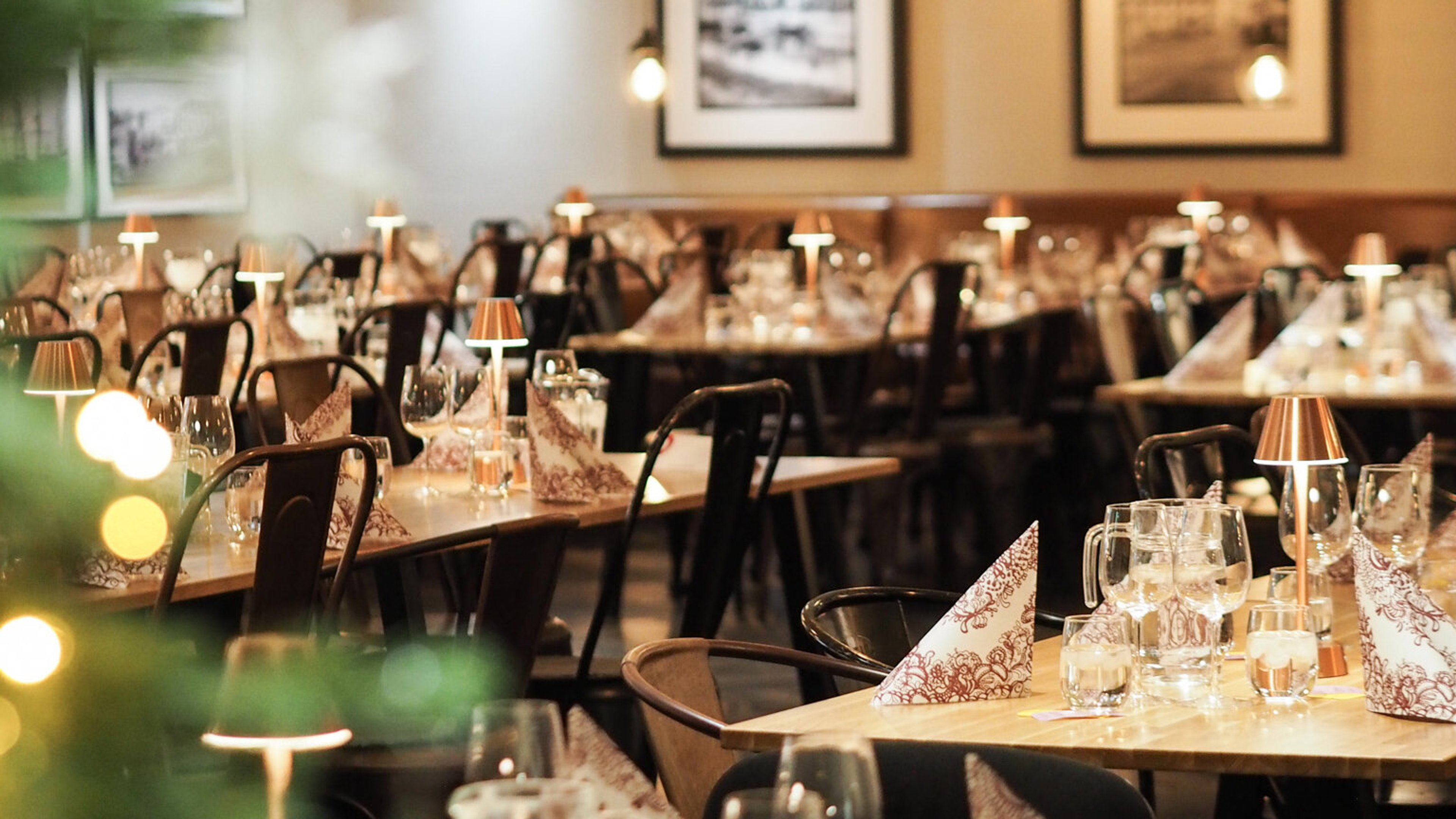 Tyylikkäästi katettu ravintolasali, jossa puiset pöydät, mustat tuolit ja pöytälamput luovat lämminhenkisen tunnelman.