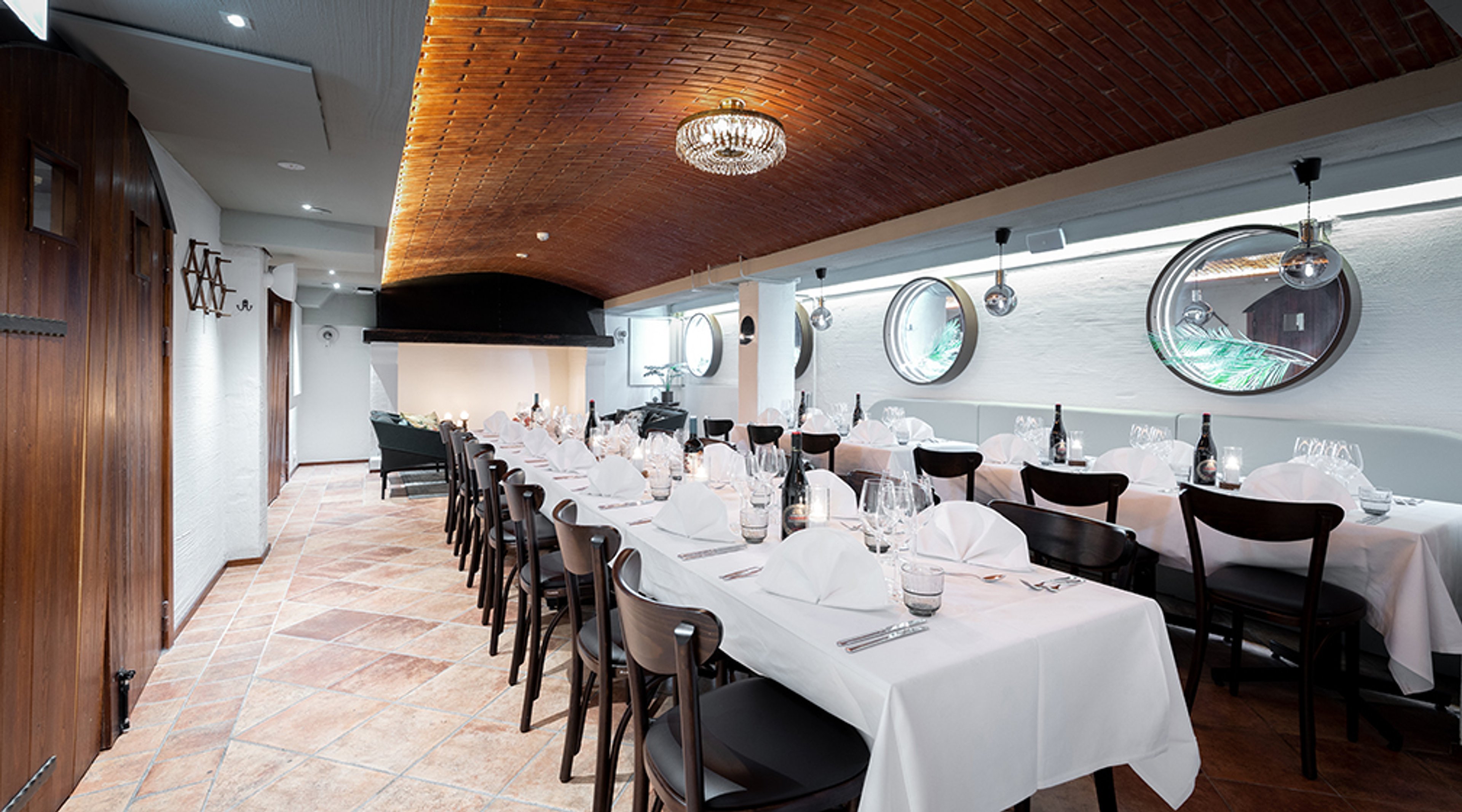 Ravintolan yksityinen kabinetti, jossa pitkä pöytä, valkoiset pöytäliinat ja kristallikruunu. Kattoa koristaa punatiili ja seinällä on pyöreitä peilejä.