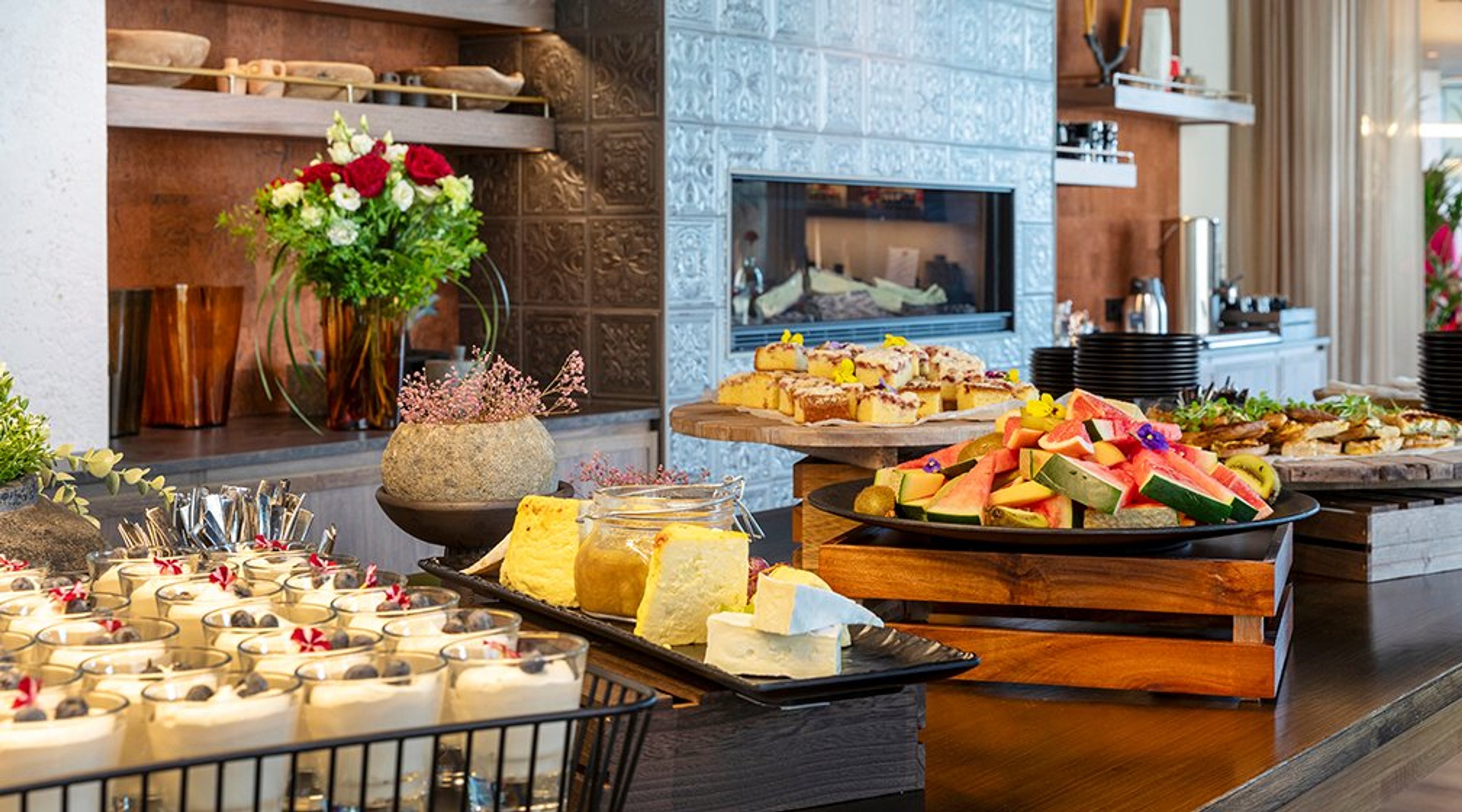 Buffetpöytä täynnä herkkuja: juustoja, hedelmiä, makeita leivoksia ja jälkiruokia. Taustalla takka ja kukka-asetelma.